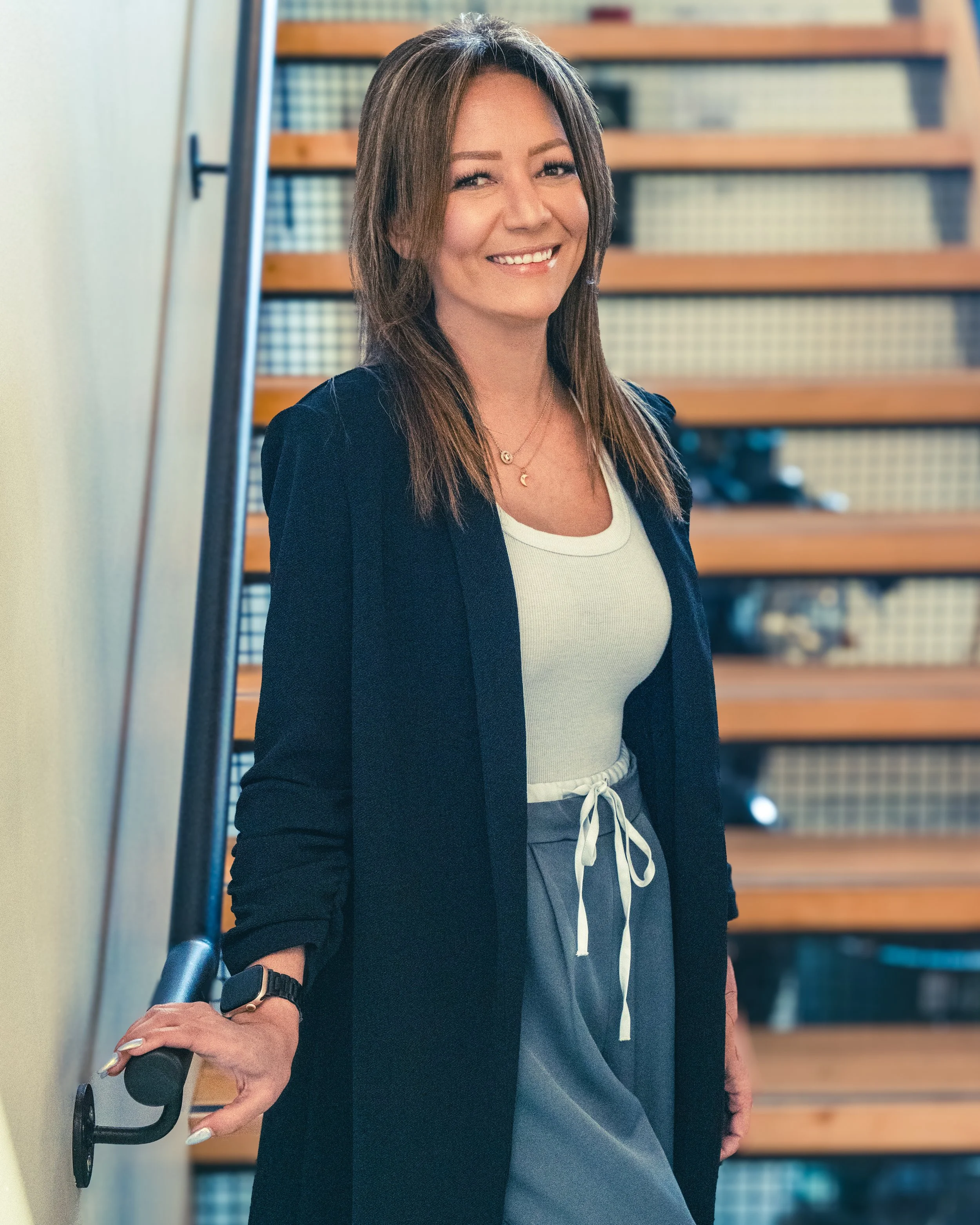 A woman with shoulder-length brown hair smiling, wearing a white top, gray pants with a white drawstring, a black blazer, and a black watch, standing on a staircase indoors.