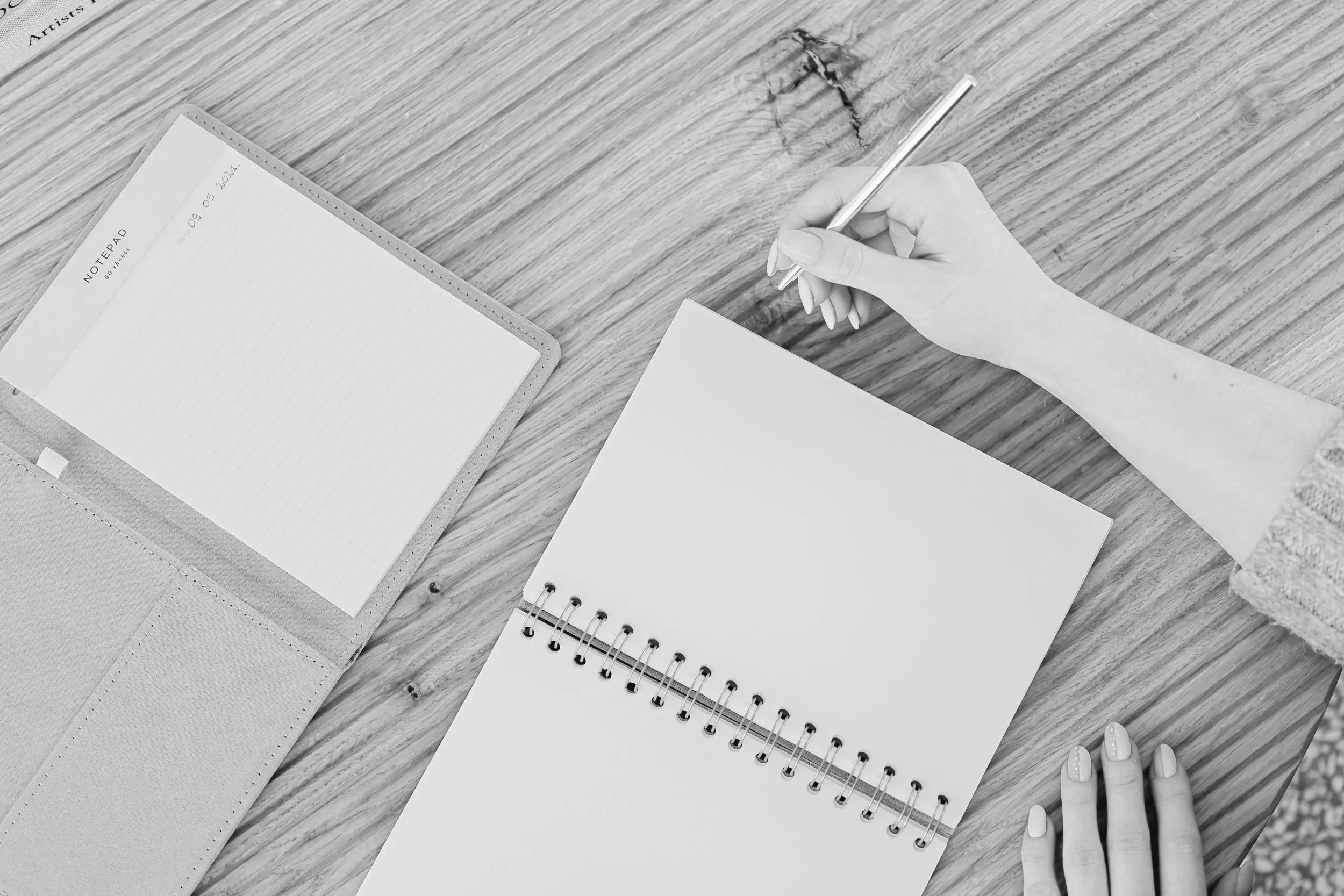 A flat lay of an open blank planner and a blank notebook with a woman's hand holding a pen.