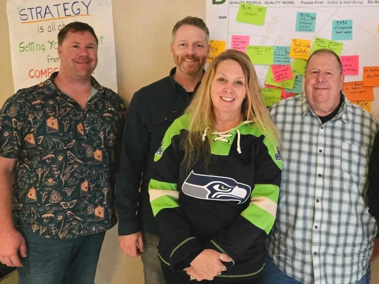 Group of four people standing together, smiling in front of a wall with colorful notes and a whiteboard with strategy notes, among them a woman in a Seattle Seahawks hockey jersey.