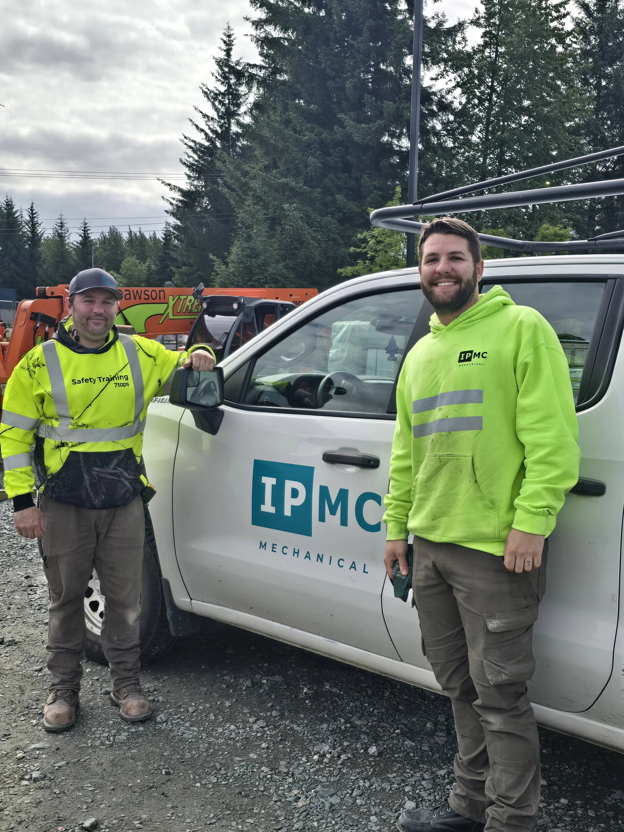 Two smiling male workers in high-visibility clothing standing outside beside a white vehicle with a company logo. There are tall trees and construction equipment in the background.