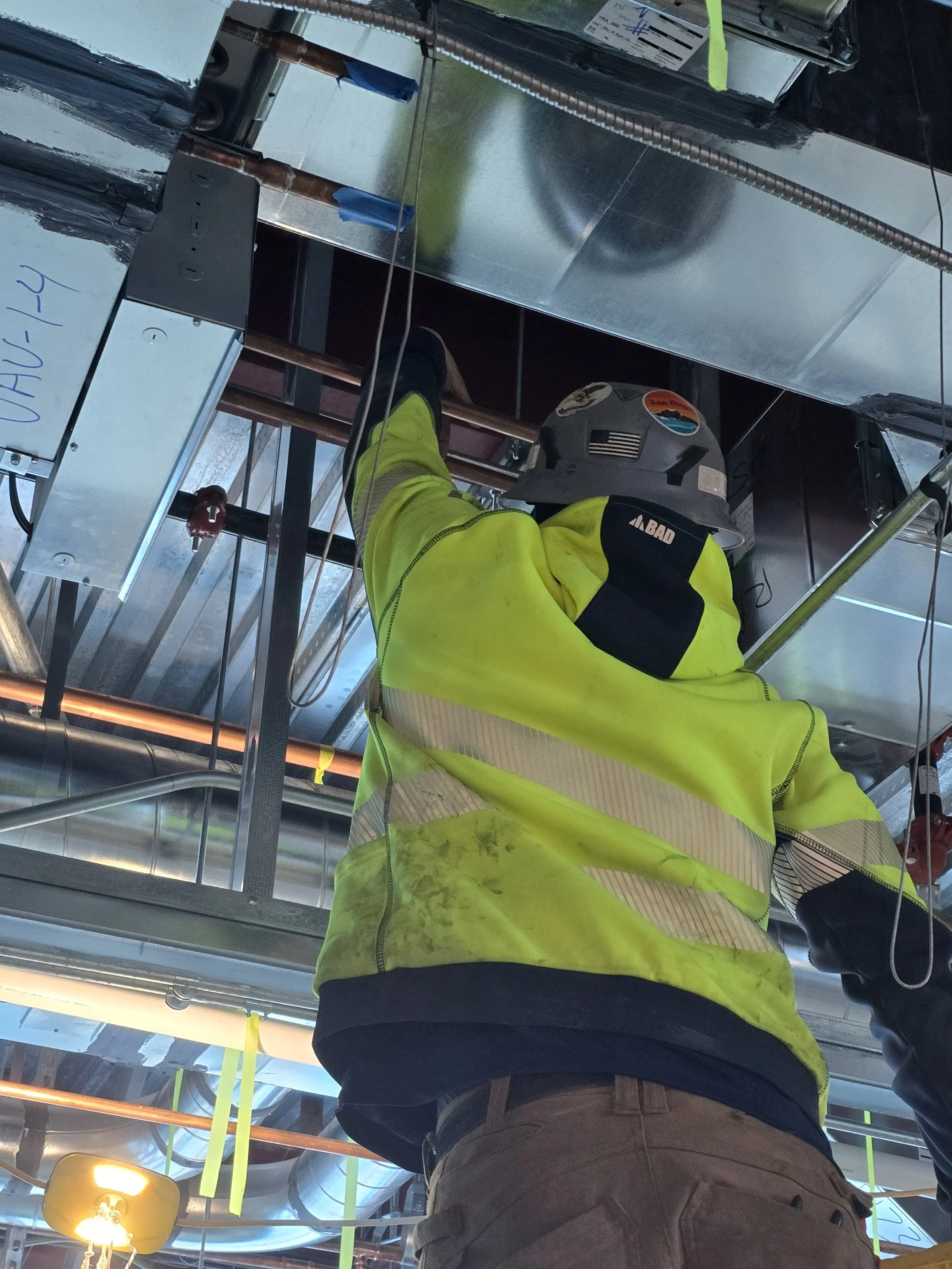 Construction worker wearing a yellow safety vest and a hard hat working on ceiling pipes and ducts in an industrial or commercial building.