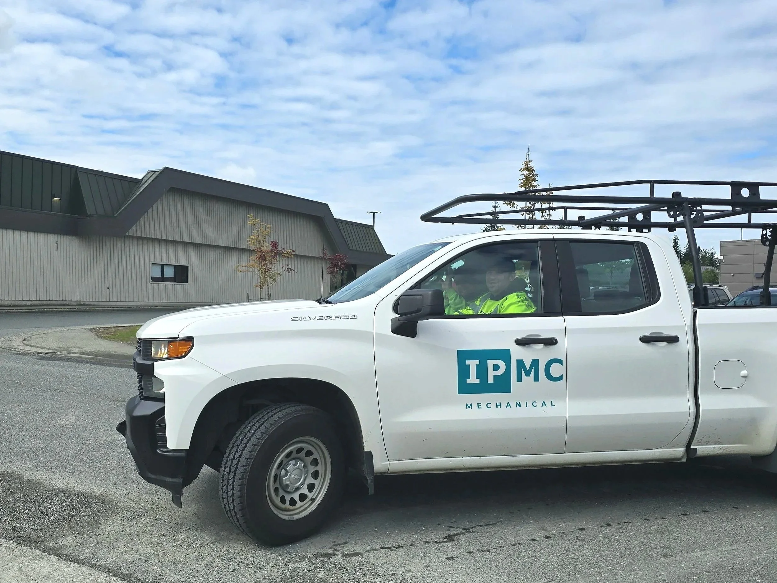 A white pickup truck with the logo 'TPMC Mechanical' on the door, parked outdoors near a building with a green roof and trees. Two people in high-visibility jackets are inside the truck.