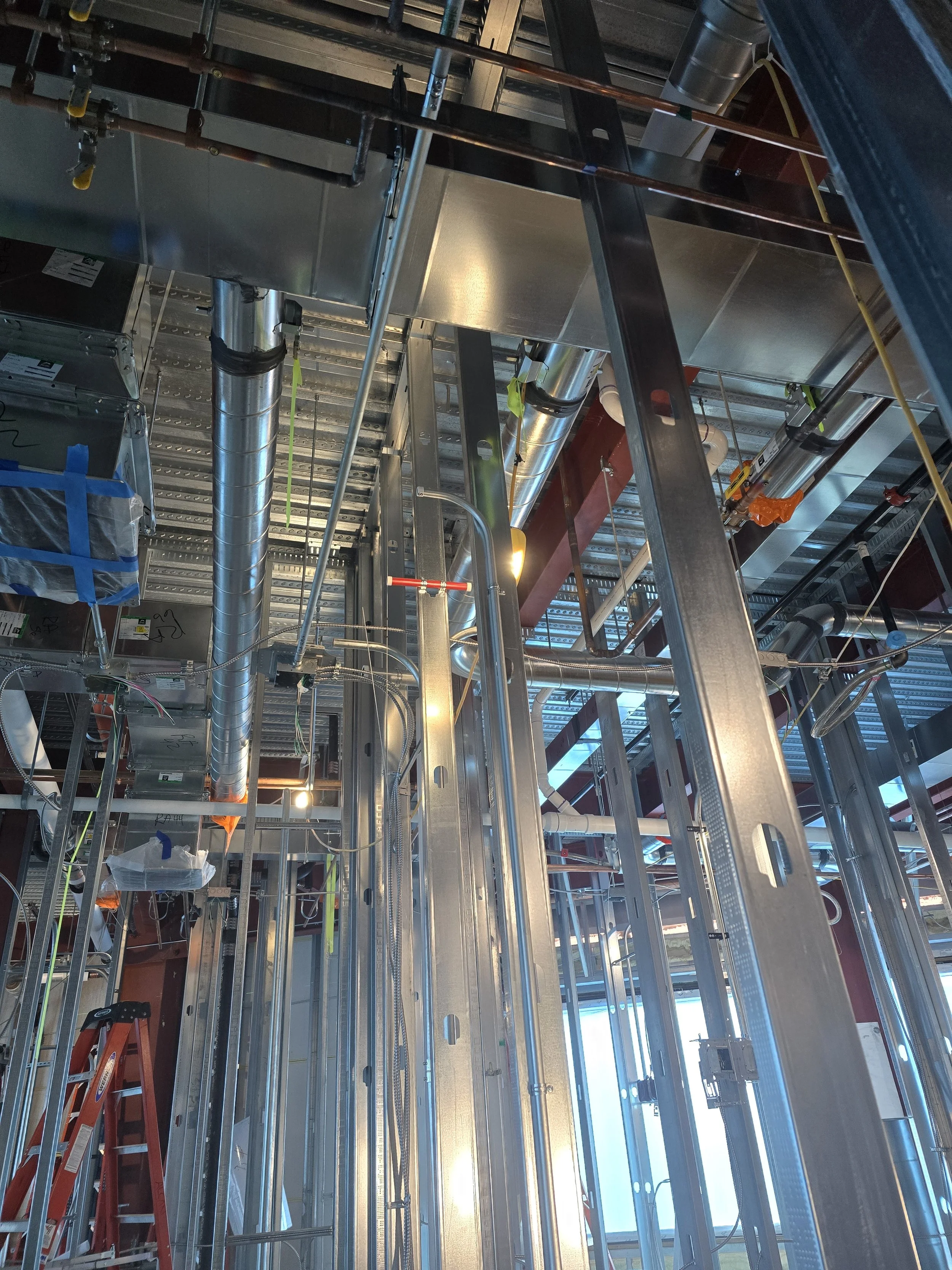Construction site interior with metal framing, pipes, and tools, including a red ladder on the left, ceiling light, and building materials.