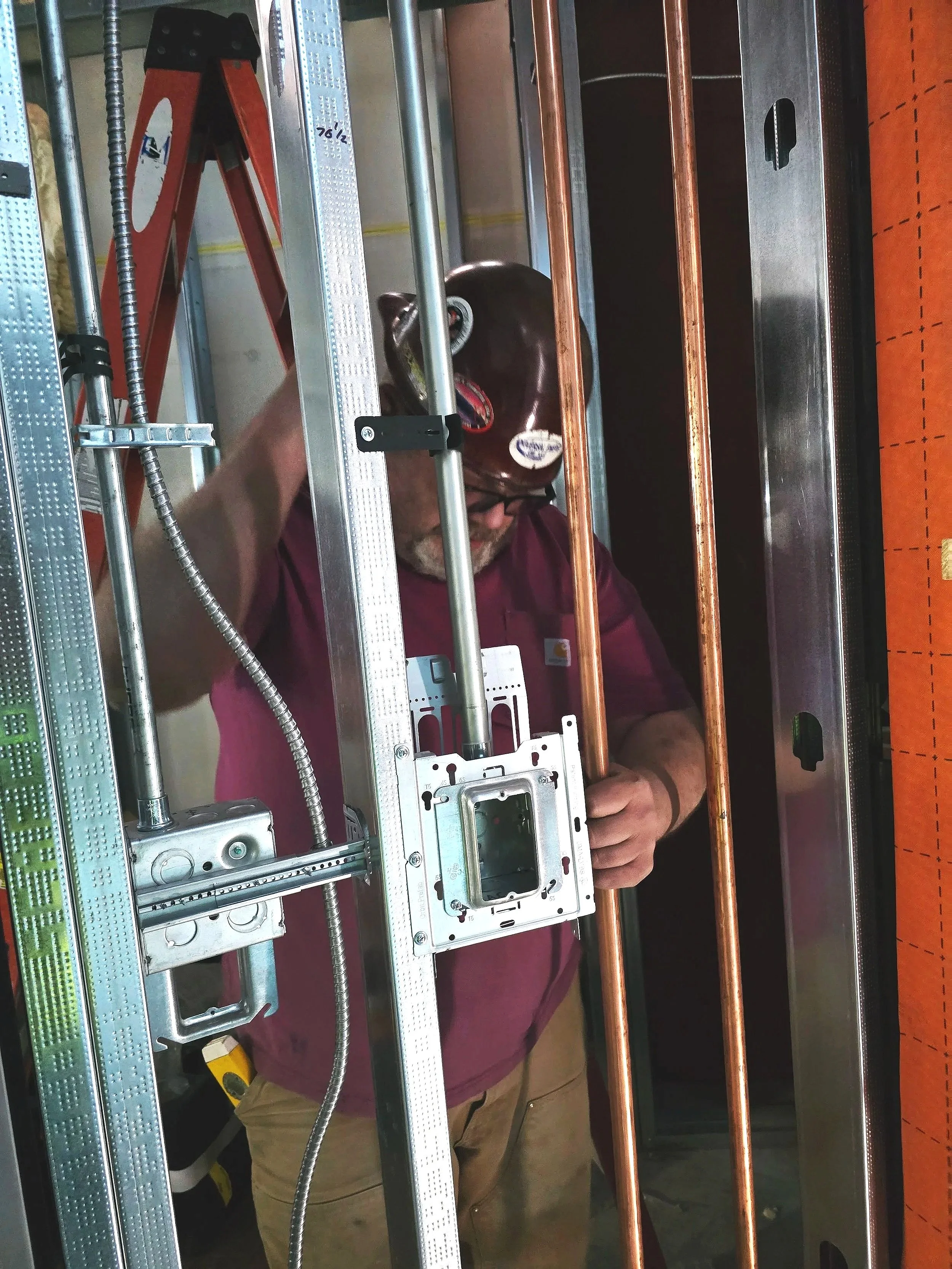 Person working on electrical or plumbing installation inside a metal frame structure, with copper pipes and electrical wiring visible.