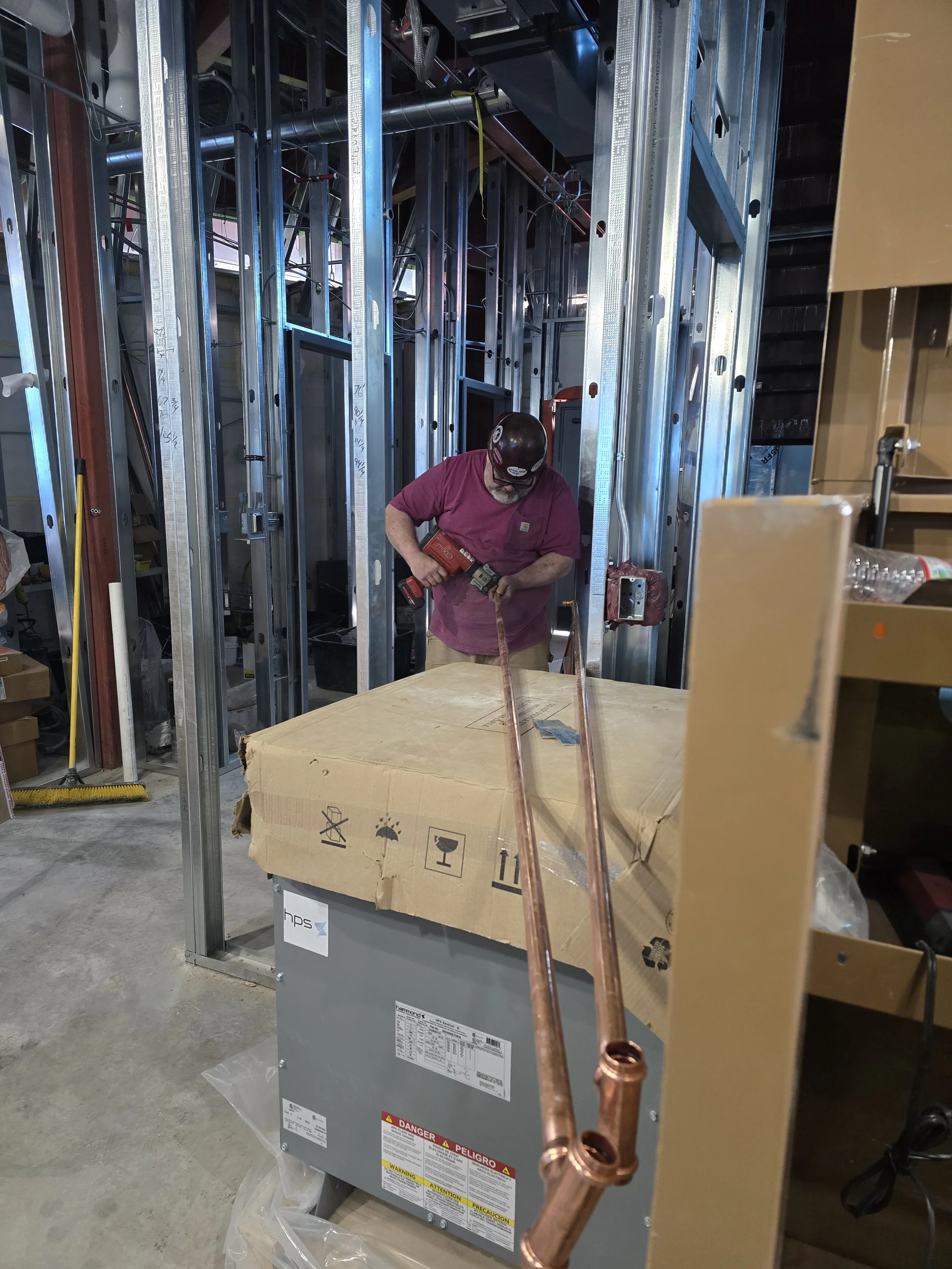 A worker in a pink shirt and sunglasses is fixing or inspecting an HVAC unit or furnace in a building under construction, with exposed metal framing and plumbing pipes in the background.