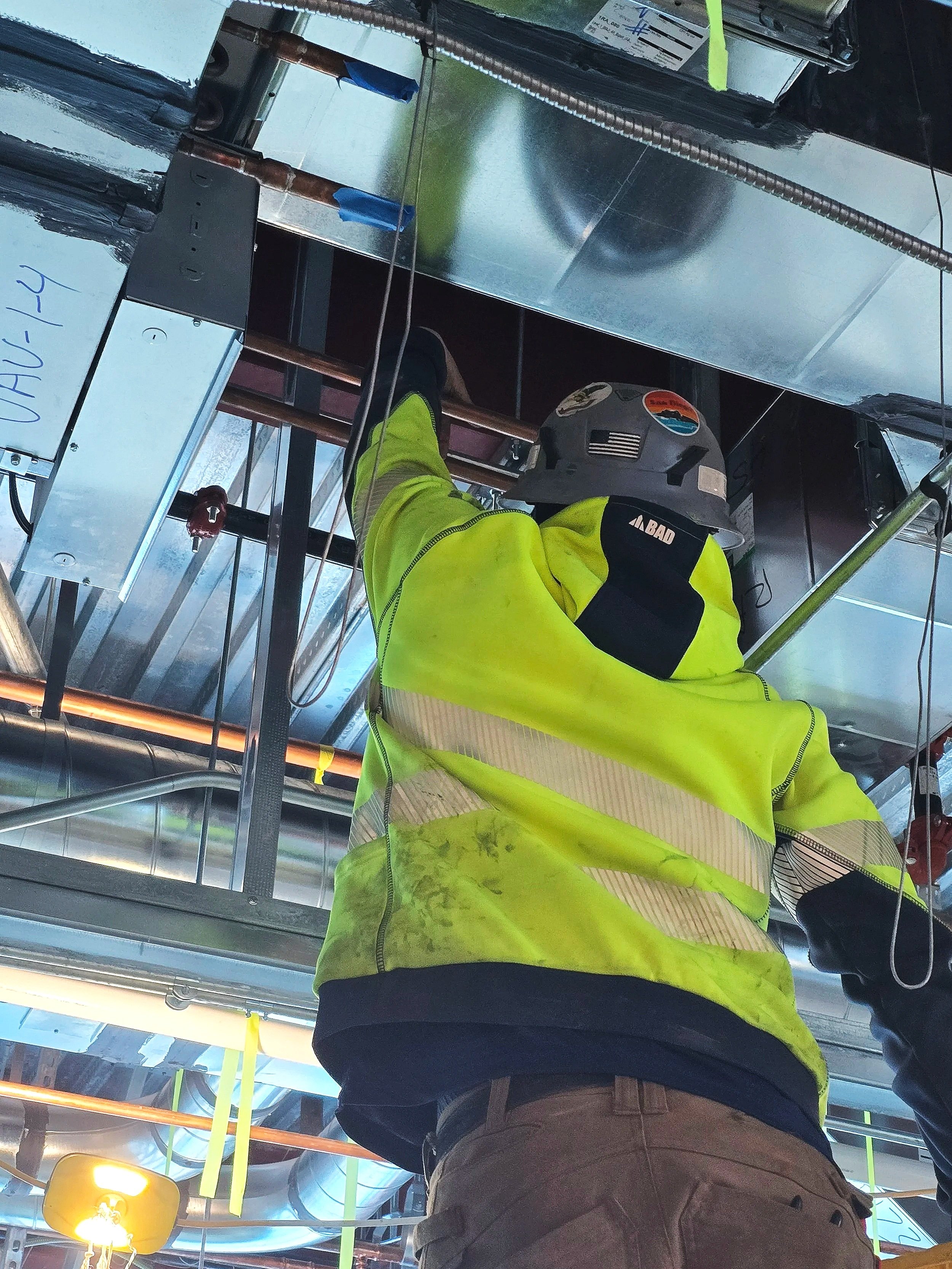 A construction worker in a yellow high-visibility jacket and helmet working on HVAC systems in a building ceiling. The worker is facing away from the camera, and the ceiling includes ducts, pipes, and electrical wiring.