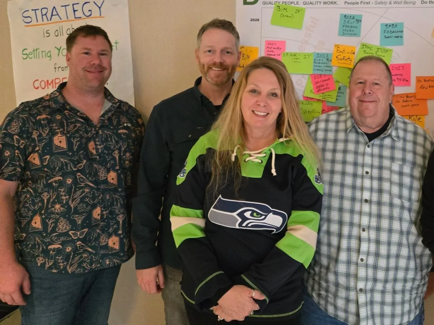 Four people standing in front of a whiteboard with colorful sticky notes. One woman is wearing a Seattle Seahawks hockey jersey, and the men are dressed casually. The background features a strategy and planning display with notes and charts.