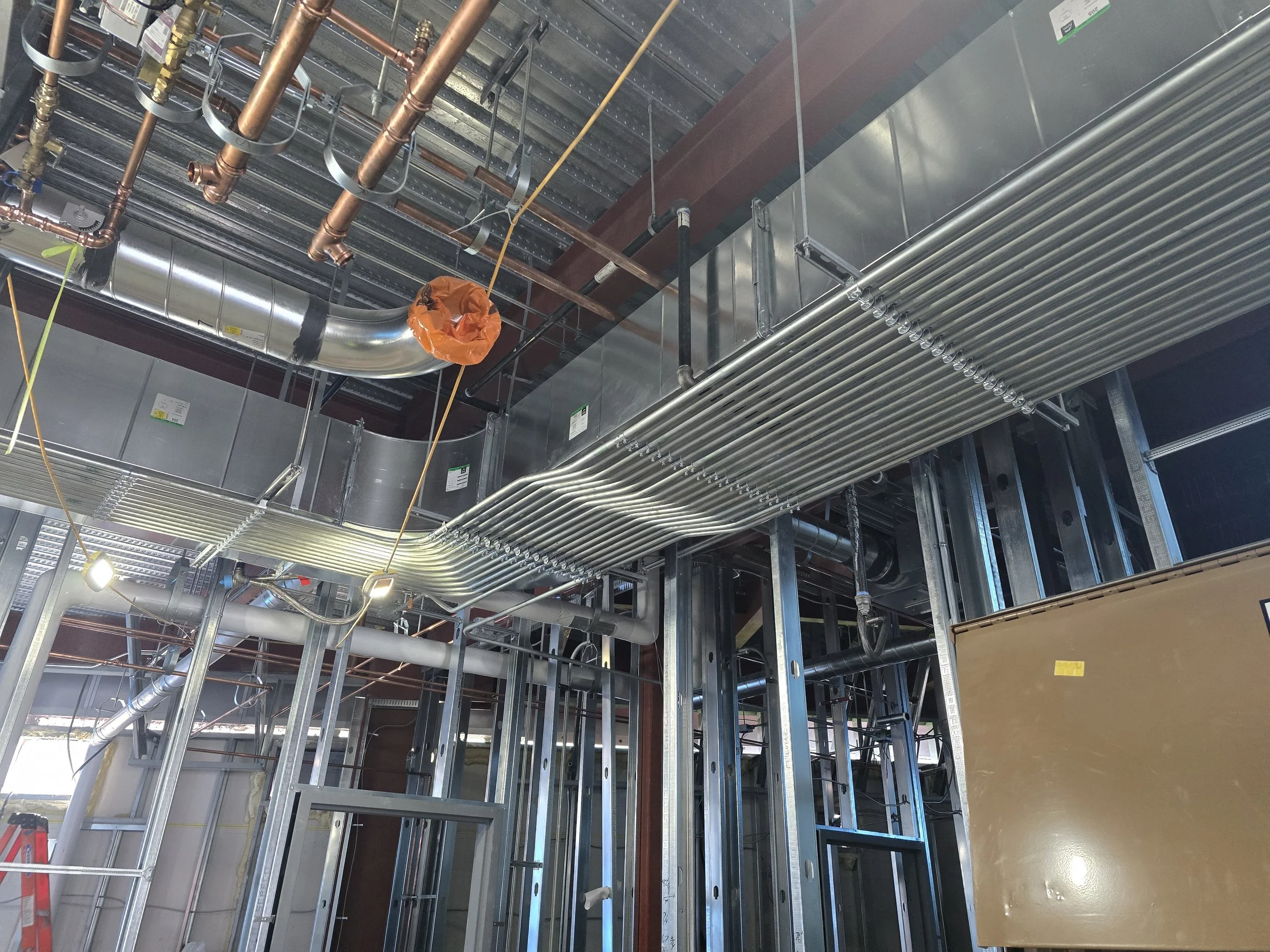 View of industrial building infrastructure with exposed copper pipes, metal ductwork, electrical conduits, and structural framing. The ceiling features a corrugated metal panel, and there's a flexible orange-covered pipe or conduit.