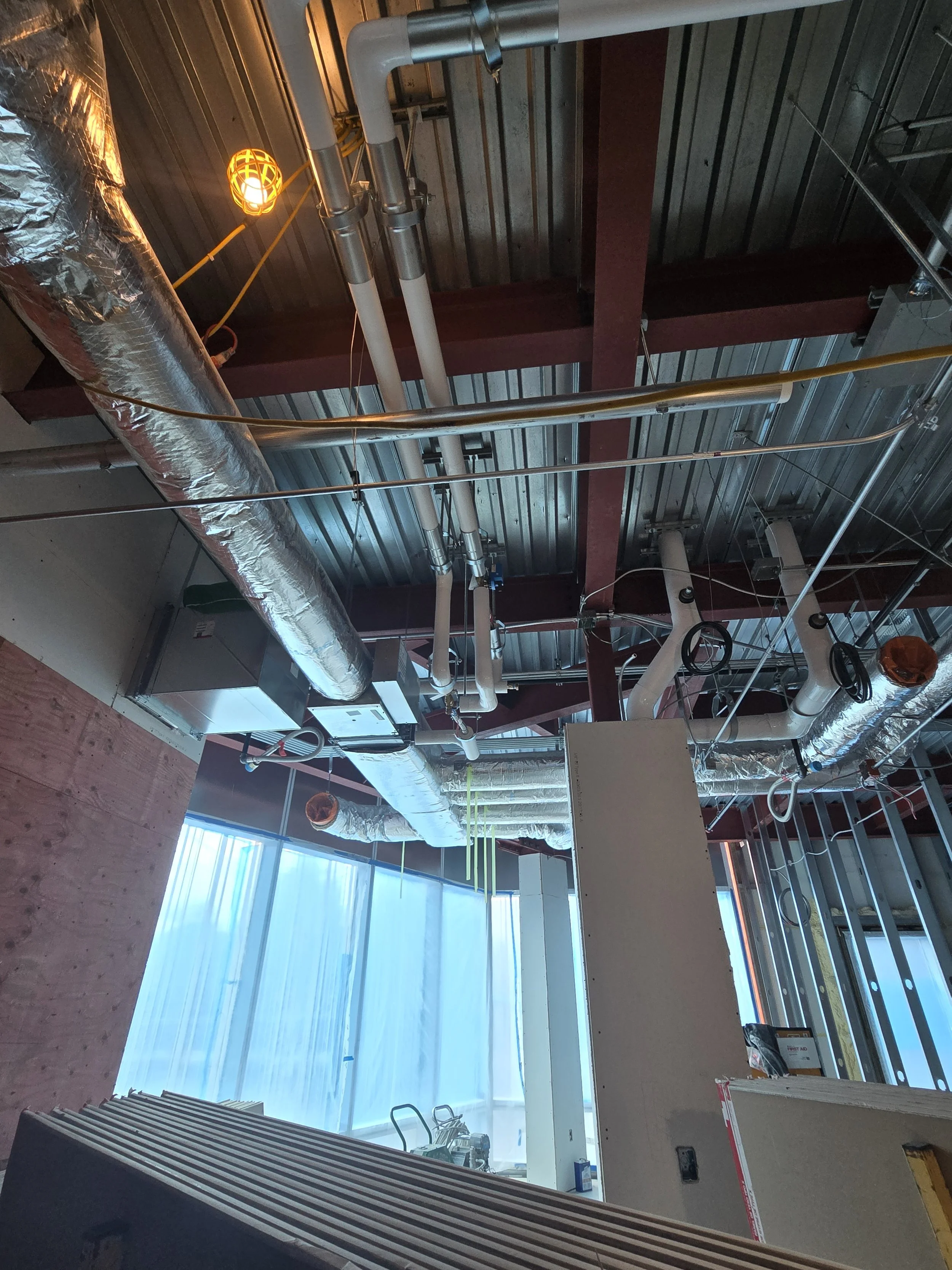 Construction site with exposed pipes, ductwork, and construction materials on the floor inside a building.