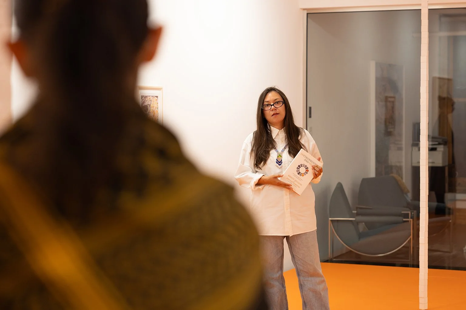 Woman with long dark hair, glasses, and a white shirt standing indoors, holding a book or pamphlet, with another person blurred in the foreground at a Dart Social Club experience.