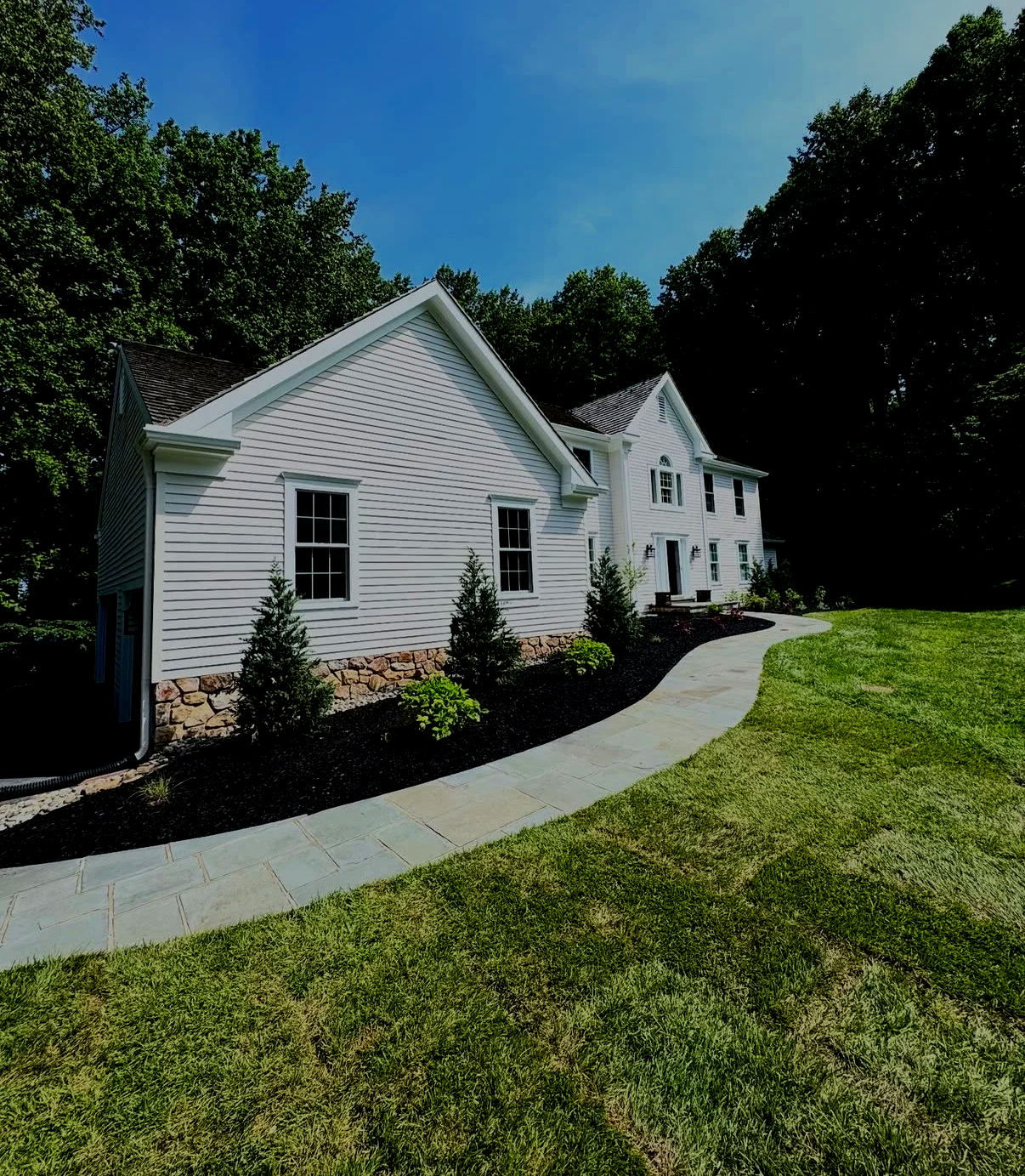 White two-story house with a stone foundation, surrounded by a curved walkway, green lawn, and trees in the background under a blue sky.