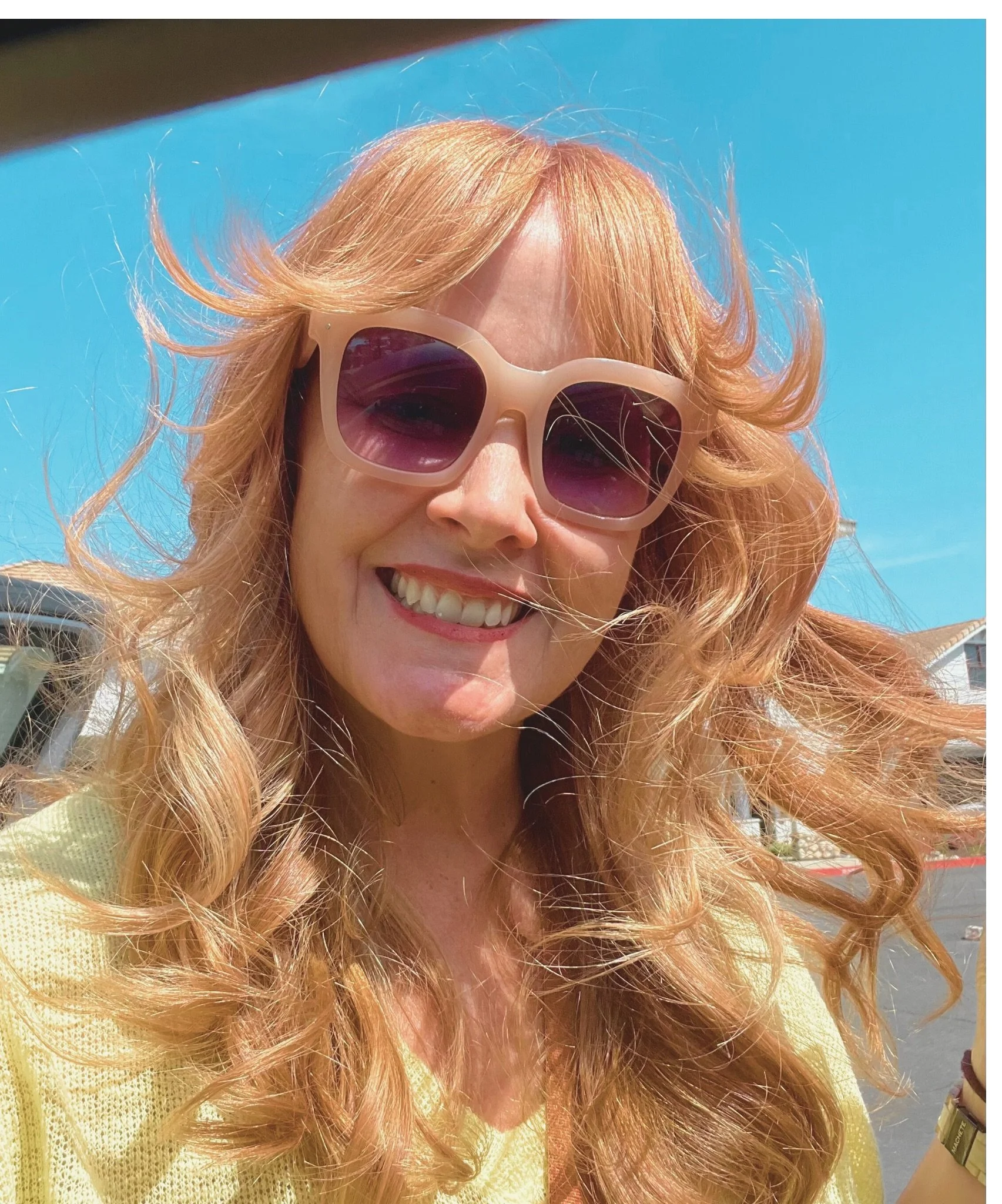 Person with long wavy hair wearing sunglasses, smiling outdoors with a clear blue sky.