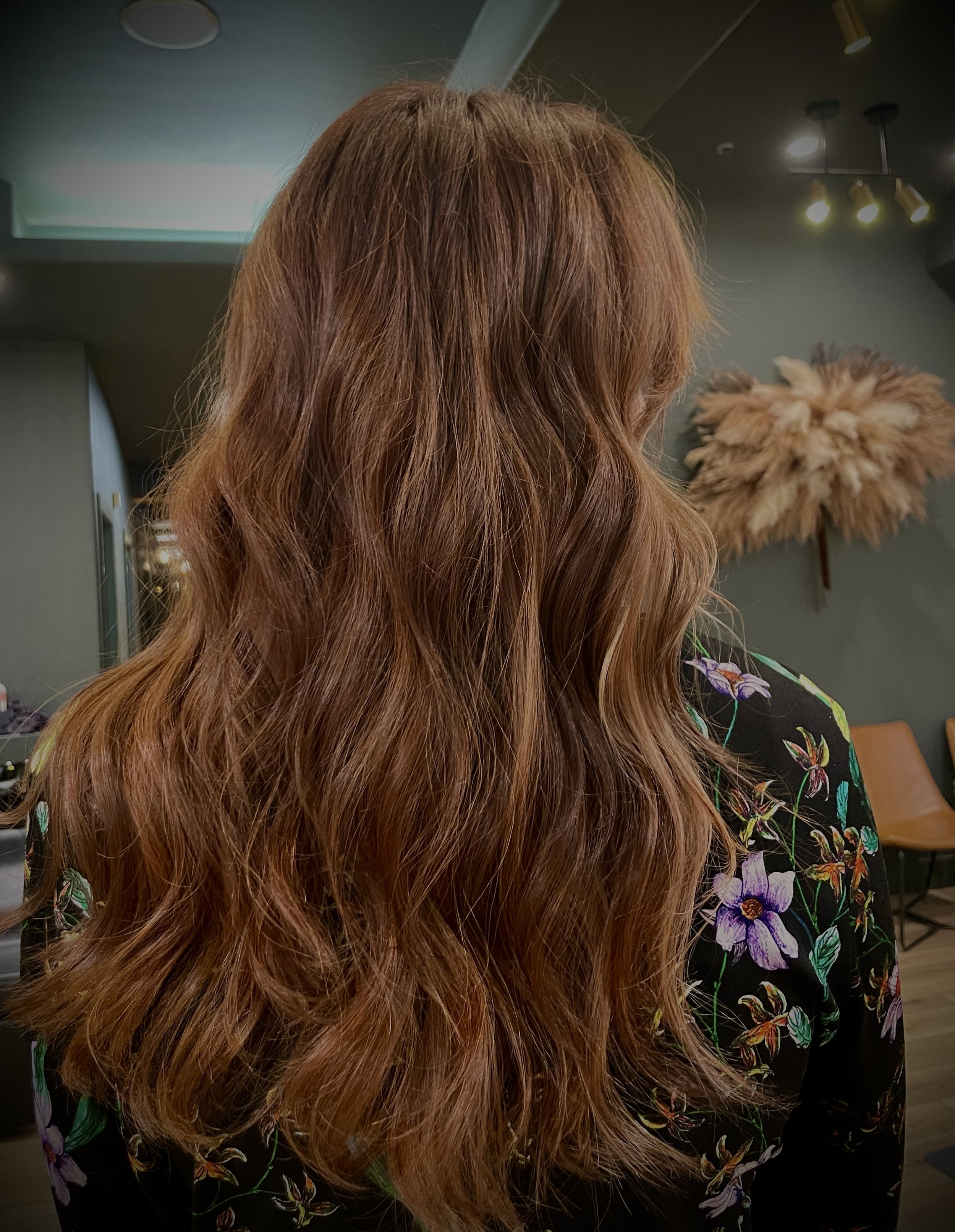 Back view of a person with long, wavy, brown hair in a dimly lit room with floral-patterned clothing and dried floral decor on the wall.