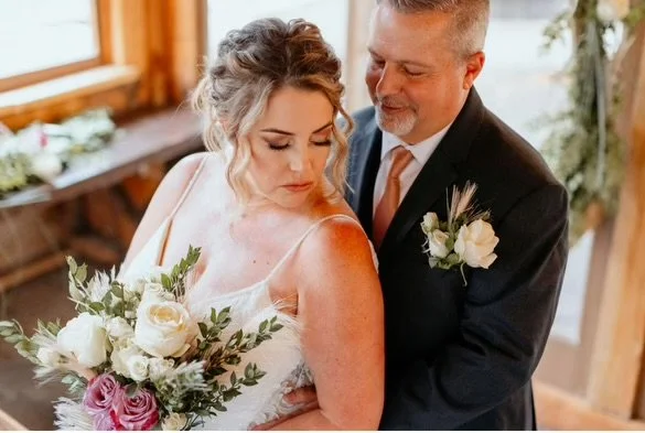 Bride and groom embracing inside a rustic venue, holding a bouquet of white and purple flowers.