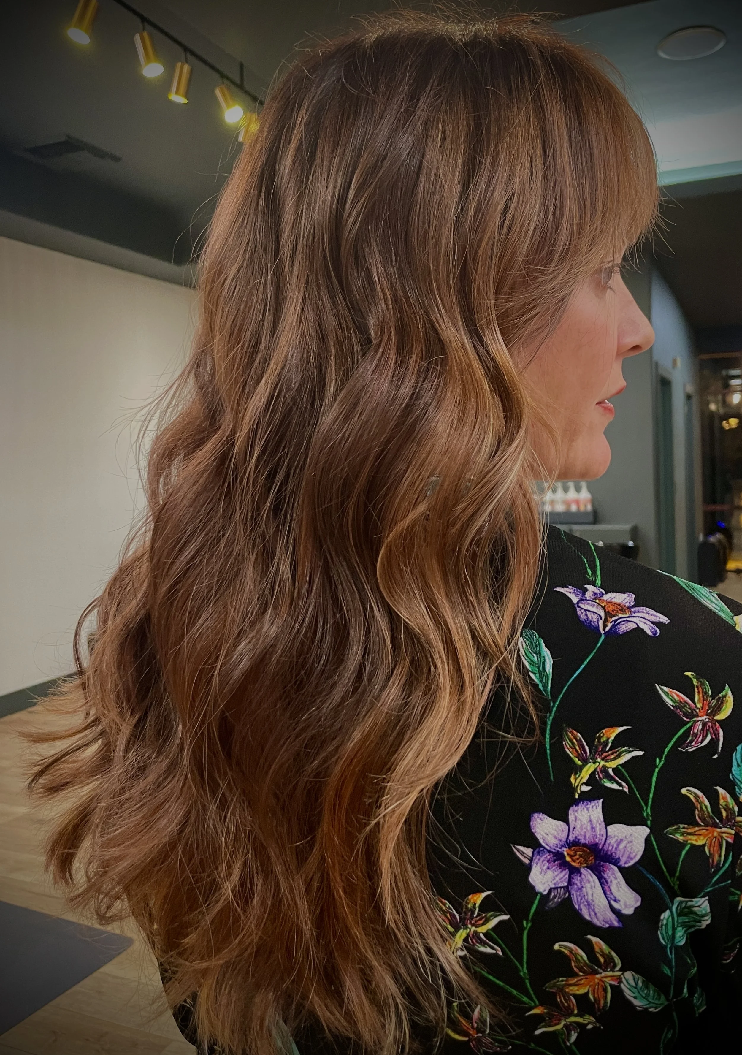 Woman with wavy brown hair in a salon, wearing a floral-patterned shirt.