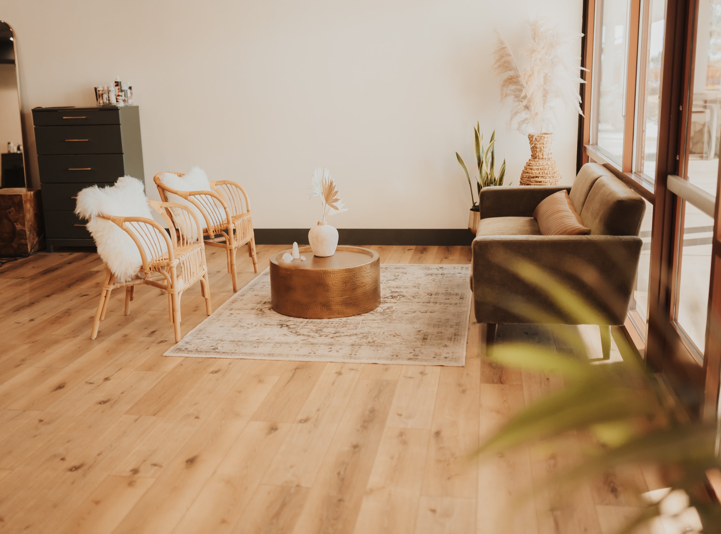 Cozy living room with wooden floor, featuring a dark green couch, two wicker chairs with white cushions, a round coffee table with a vase, and plants by a window; a cabinet in the background.