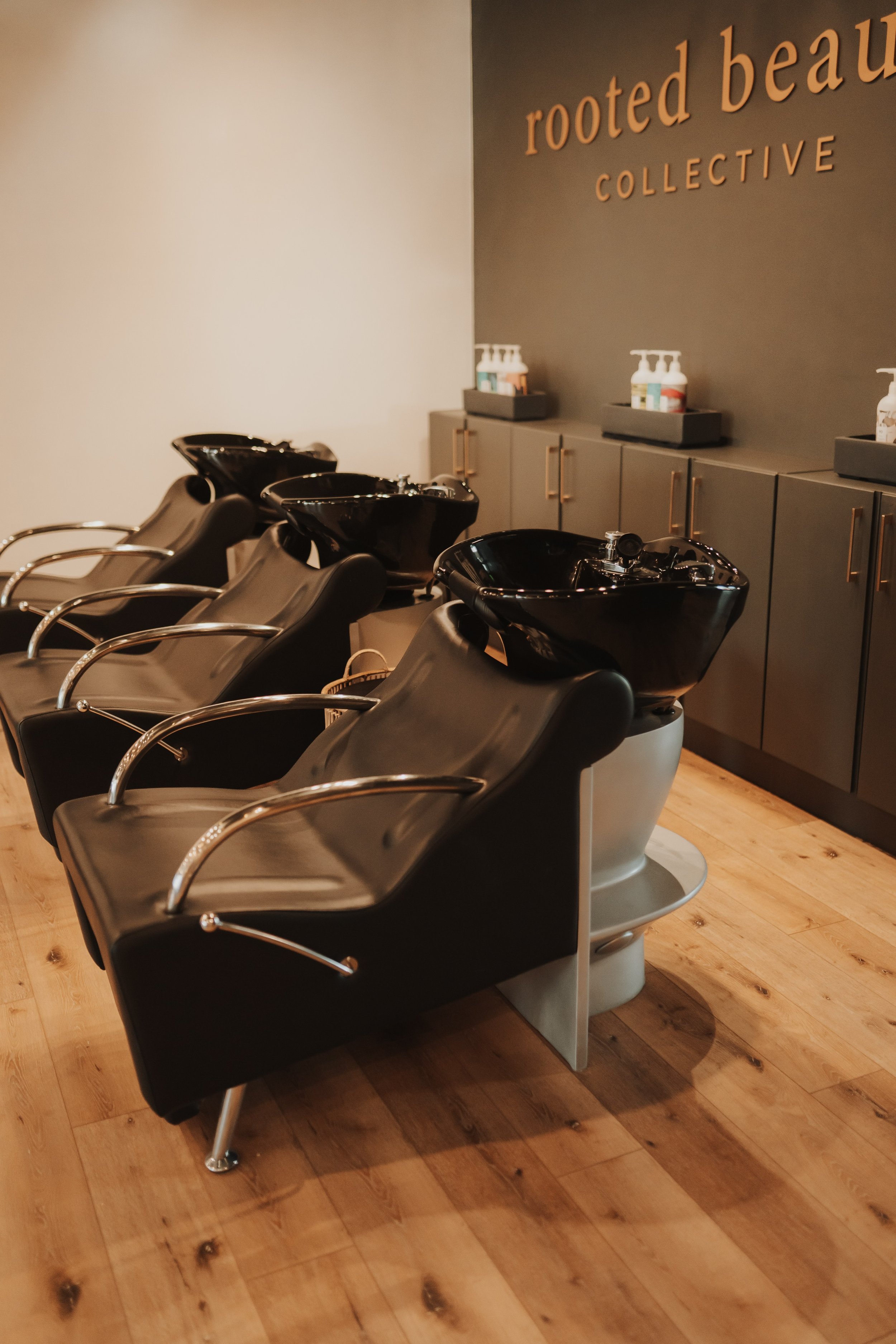 Salon with black wash chairs and shampoo sinks, wooden floor, products on cabinets, "rooted beauty COLLECTIVE" sign on wall.