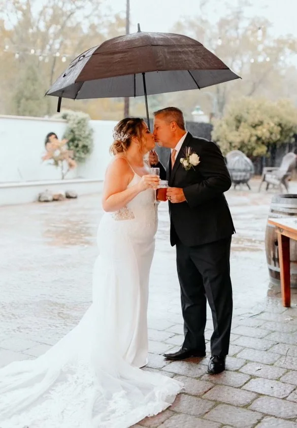 A bride and groom kissing under a black umbrella on a rainy day. The bride is wearing a white wedding dress, and the groom is in a black suit with a white boutonniere. They are holding drinks and standing on a wet brick patio.