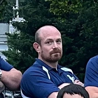 A man with a bald head and beard wearing a blue collared shirt with white accents, standing outdoors with trees in the background.