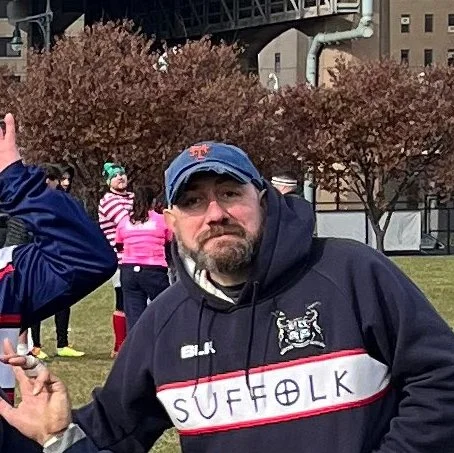 A man wearing a baseball cap and a hoodie with 'SUFFOLK' written on it, standing outdoors with trees and people in the background.