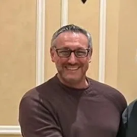 A smiling man with glasses and short gray hair, wearing a brown long-sleeve shirt, standing indoors with beige walls and white wooden trim.