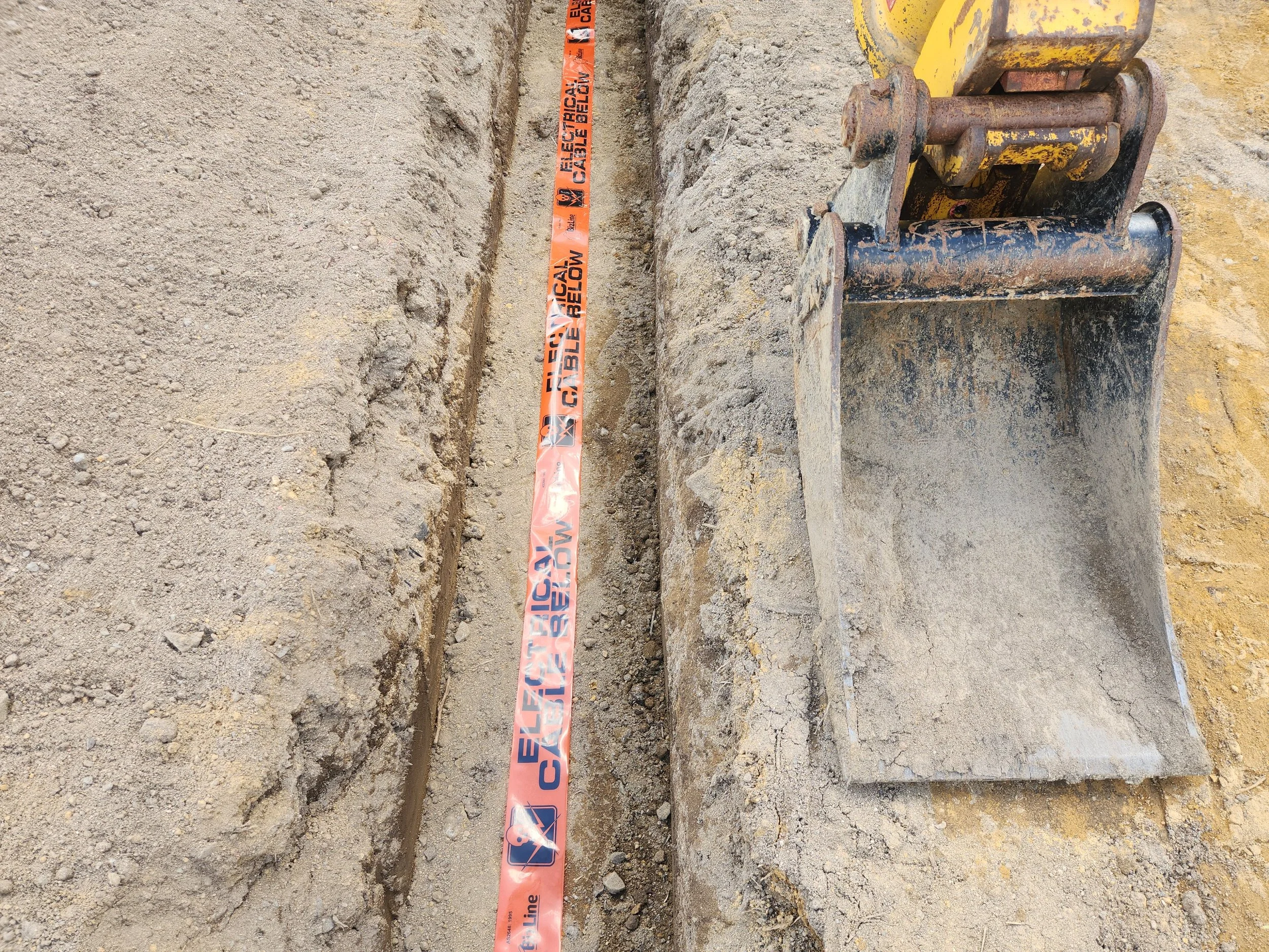 danger tape being laid while an excavator trenches for an electrical main cable
