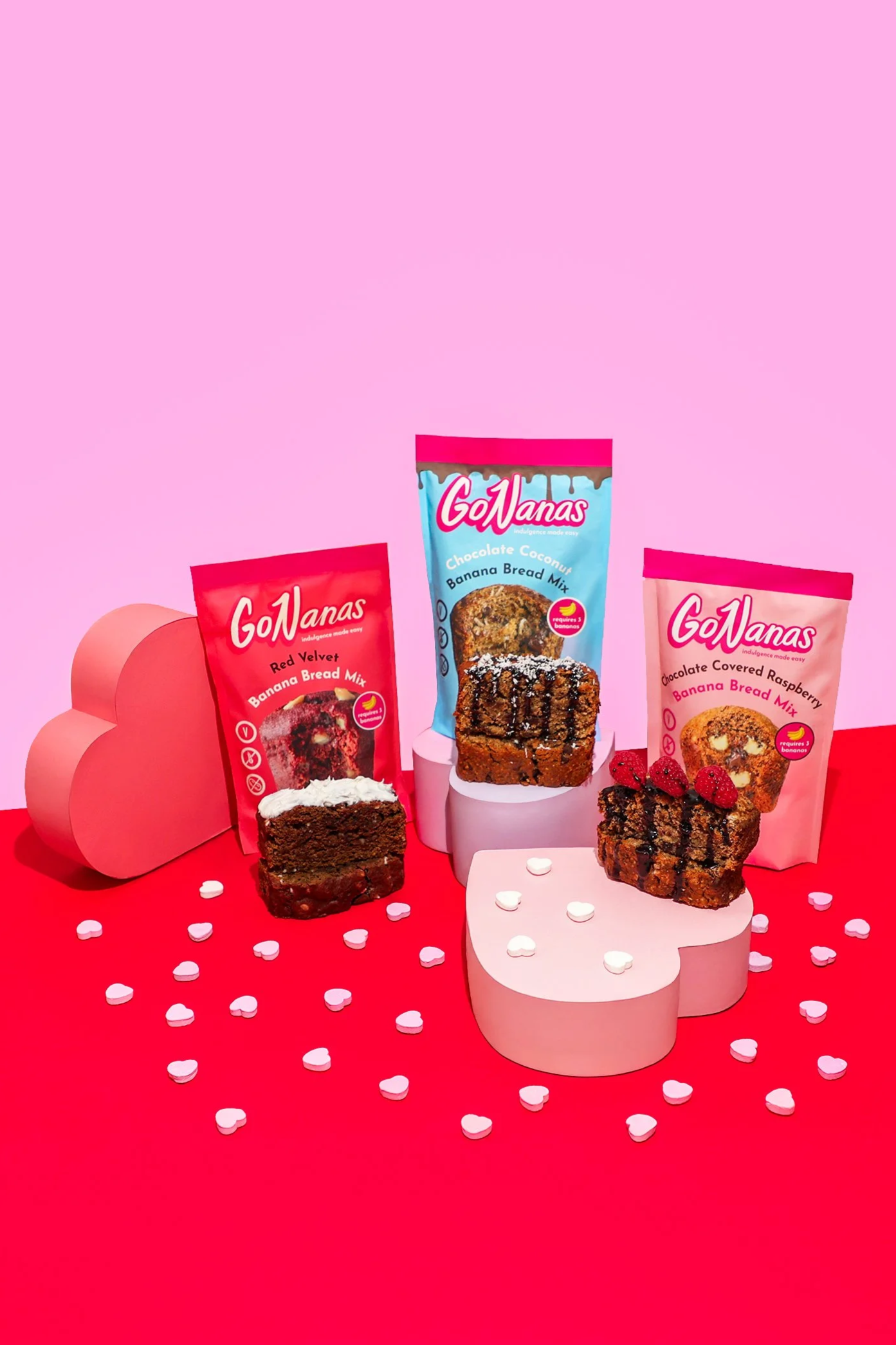Display of assorted GoNanas banana bread mixes with decorative pink heart-shaped platforms and scattered pink heart-shaped confetti on a pink and red background. For Valentine's Day banana bread launch. Colorful commercial product photography.