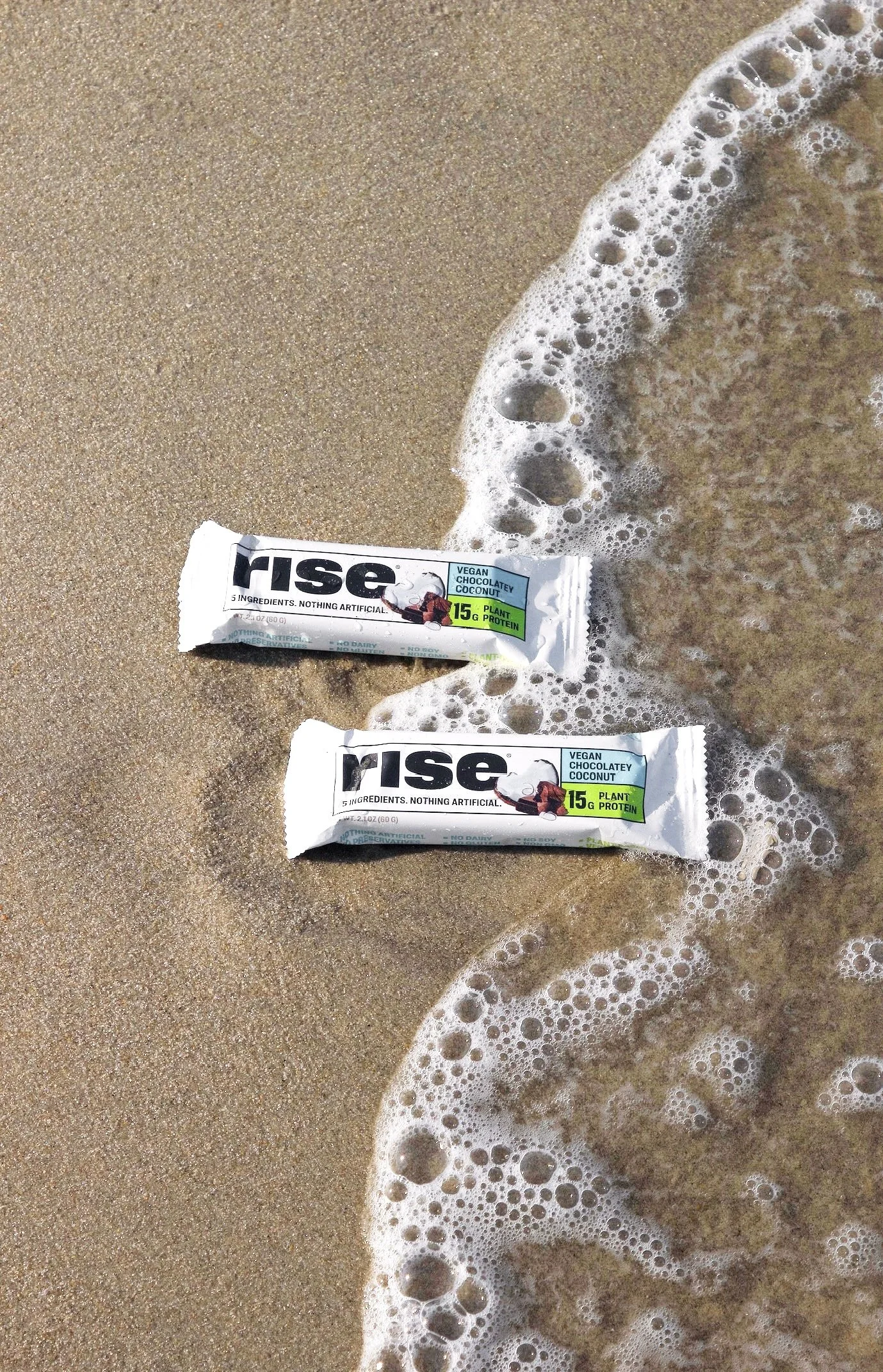 Two Rise energy bars on sandy beach with ocean waves washing over them. Commercial product photography for CPG food, and beverage brands, creating social media, and marketing-ready product images. Natural lifestyle beach photography studio.