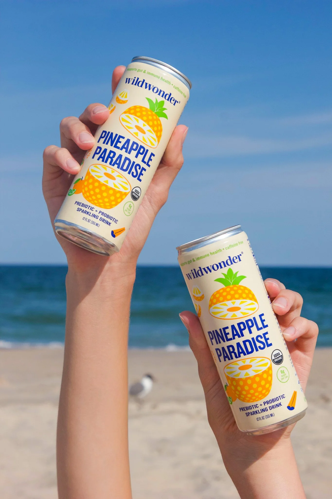Two hands holding cans of Wild Wonder Pineapple Paradise sparkling drink on a beach. Commercial product photography for CPG, food, and beverage brands, creating e-commerce, social media, and marketing-ready product images. Lifestyle model photos.