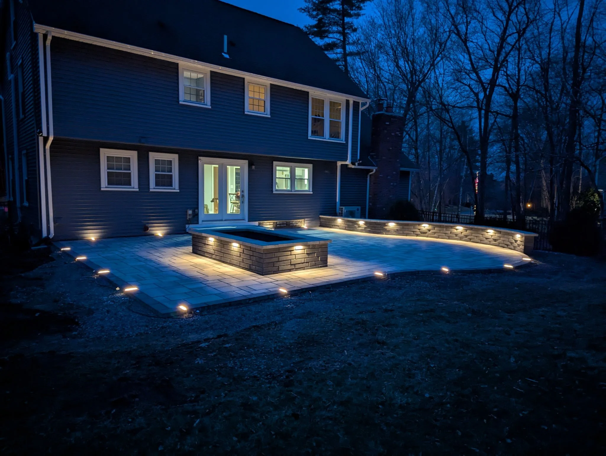 Backyard patio with brick fire pit and paver patio, illuminated by landscape lighting at dusk, adjacent to a two-story house with light inside and surrounded by trees.