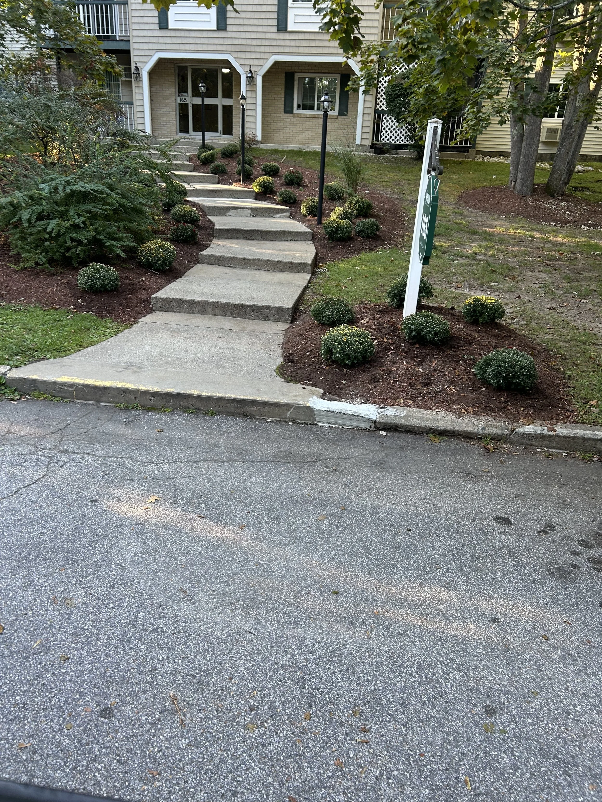 Concrete sidewalk  in Manchester NH leading up to a house entrance with landscaped bushes and a sign on the lawn.