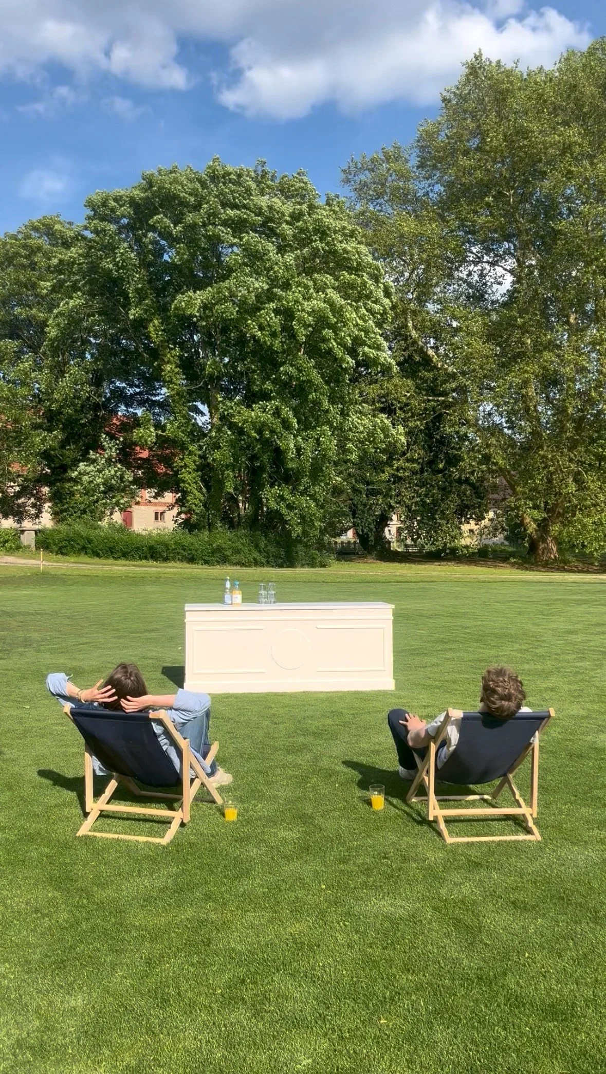 Two children sitting on lounge chairs on a grassy lawn, facing a white bar with drinks on top, with green trees and a blue sky in the background.