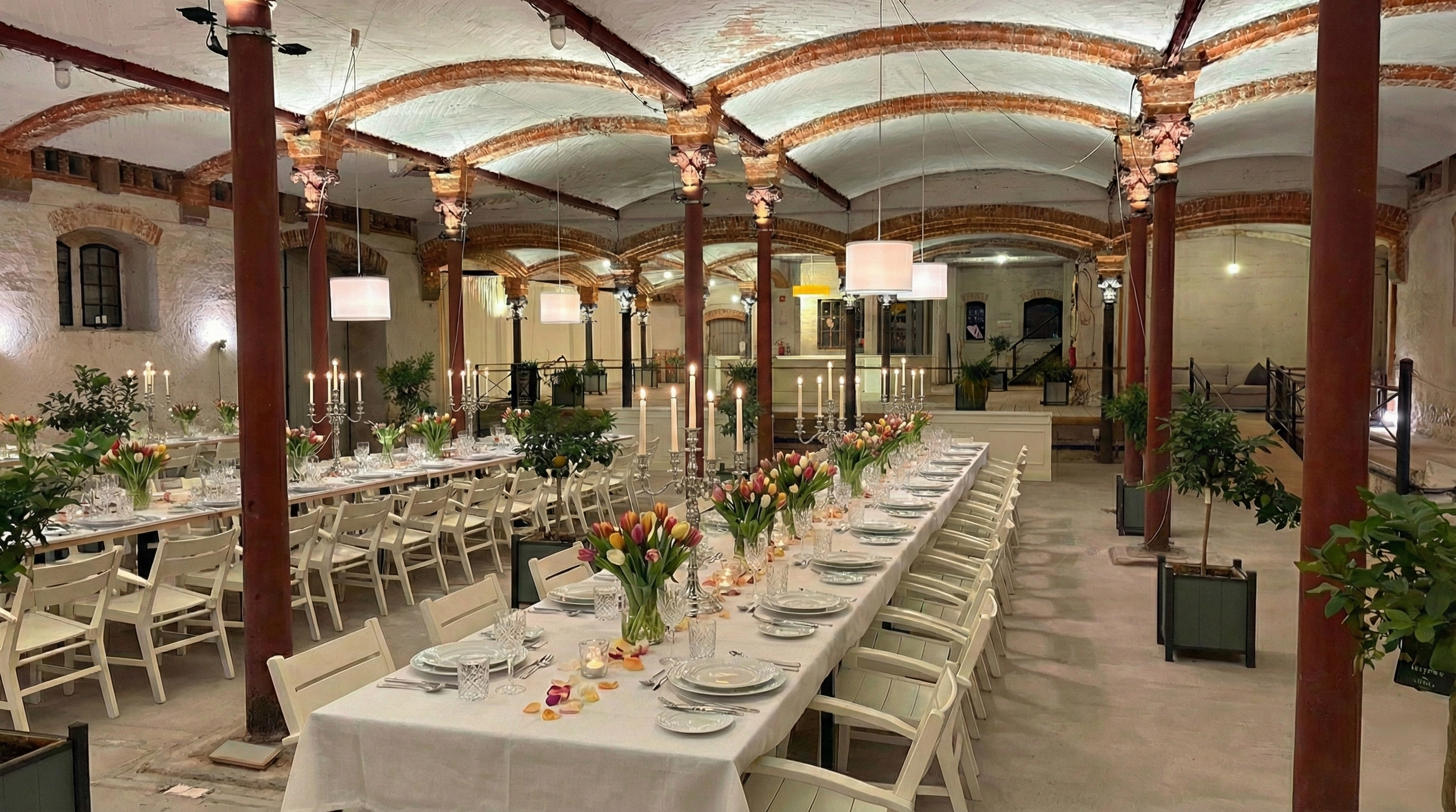 Elegant dining setup with long tables covered in white tablecloths, decorated with tulip flower arrangements, candelabras, and candles. The room has an arched ceiling with exposed brick and columns, soft lighting, and potted plants along the sides.