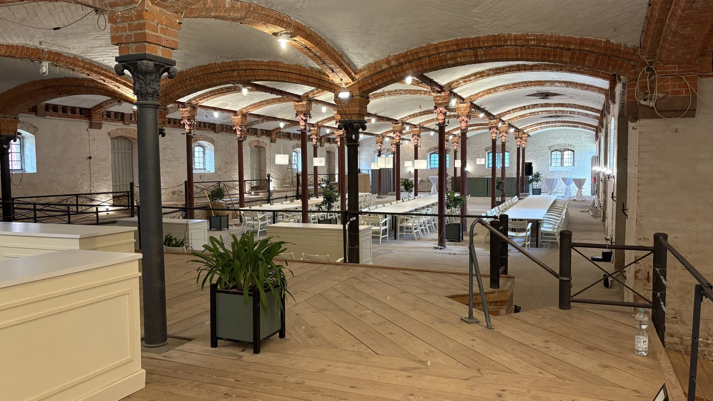 Interior of a spacious event hall with brick arches, wooden flooring, long tables with white chairs, potted plants, and modern lighting fixtures.