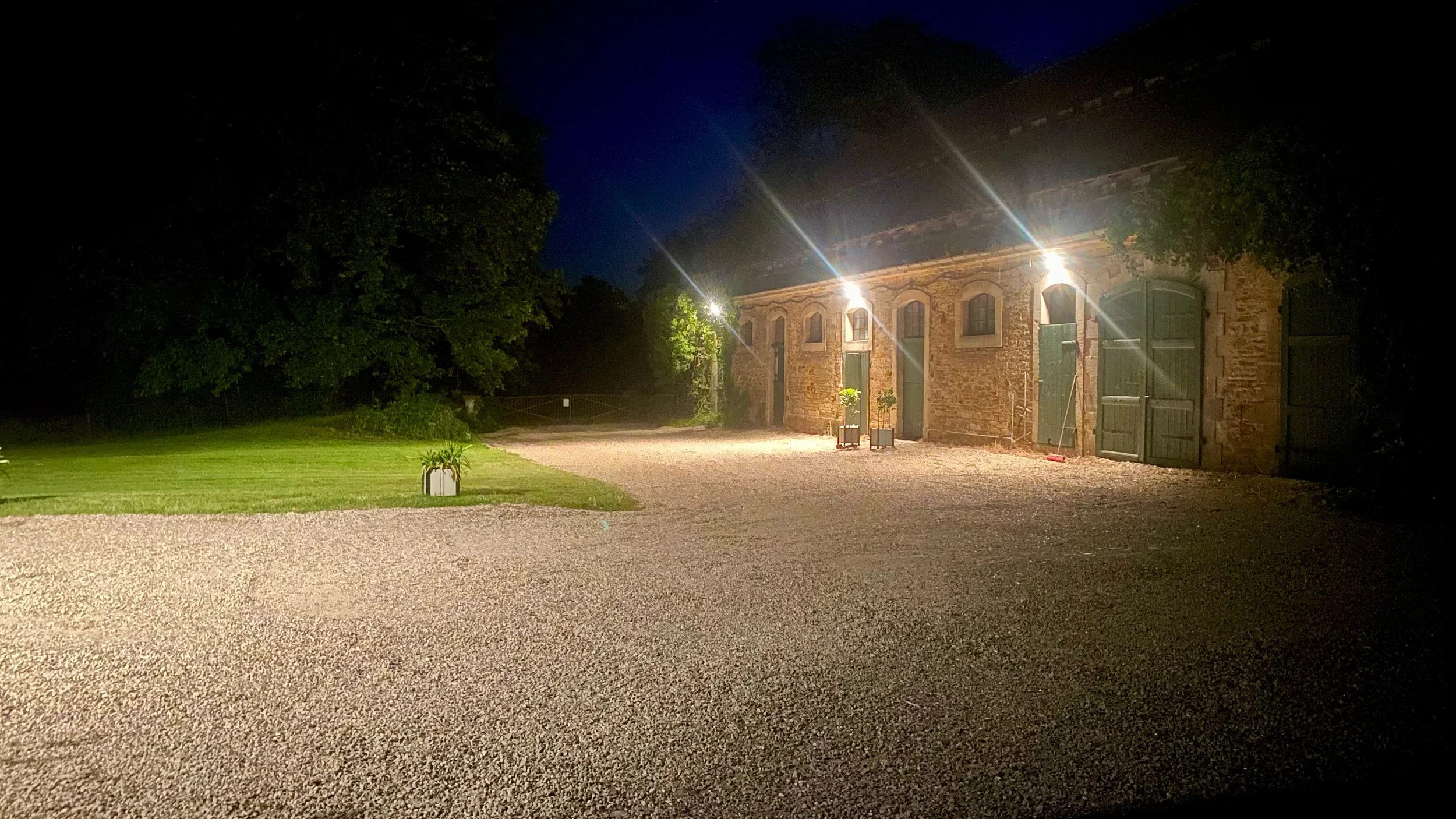 A nighttime scene of a rustic brick building with large green doors and small windows, illuminated by exterior lights. There is a gravel area in front and some potted plants nearby, with trees in the background.