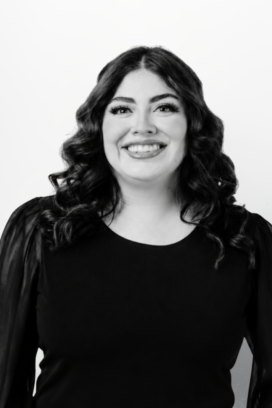 Black and white portrait of a woman with long dark wavy hair, smiling, wearing a black top and a black jacket.