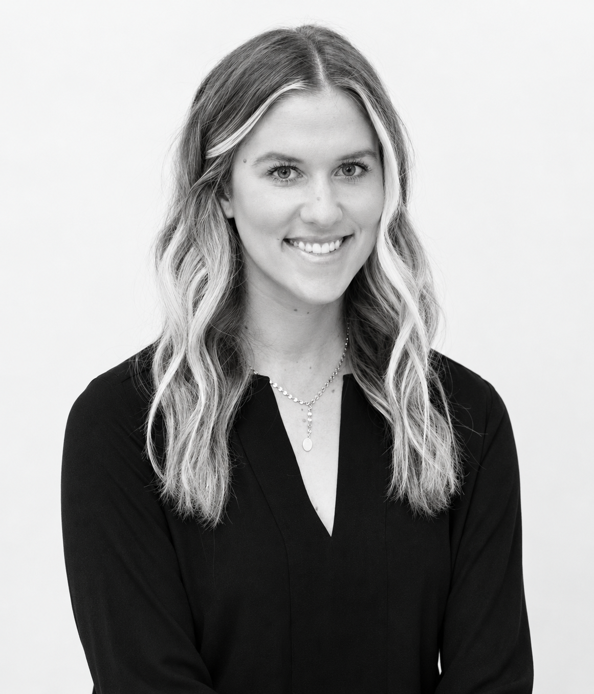 Black and white portrait of a woman with wavy hair, smiling, wearing a necklace and a dark top, standing against a plain light background.