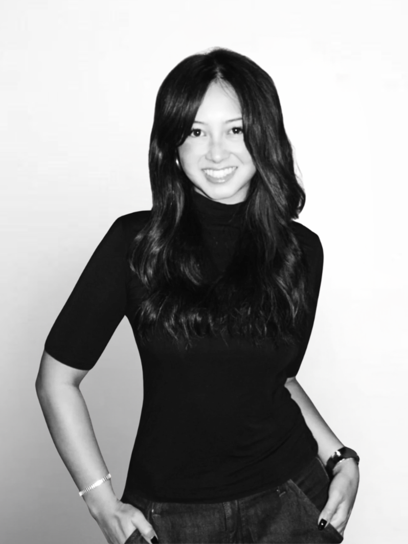 Black and white photo of a young woman with long, wavy hair, smiling, wearing a black turtleneck and jeans, with her hands in her pockets.