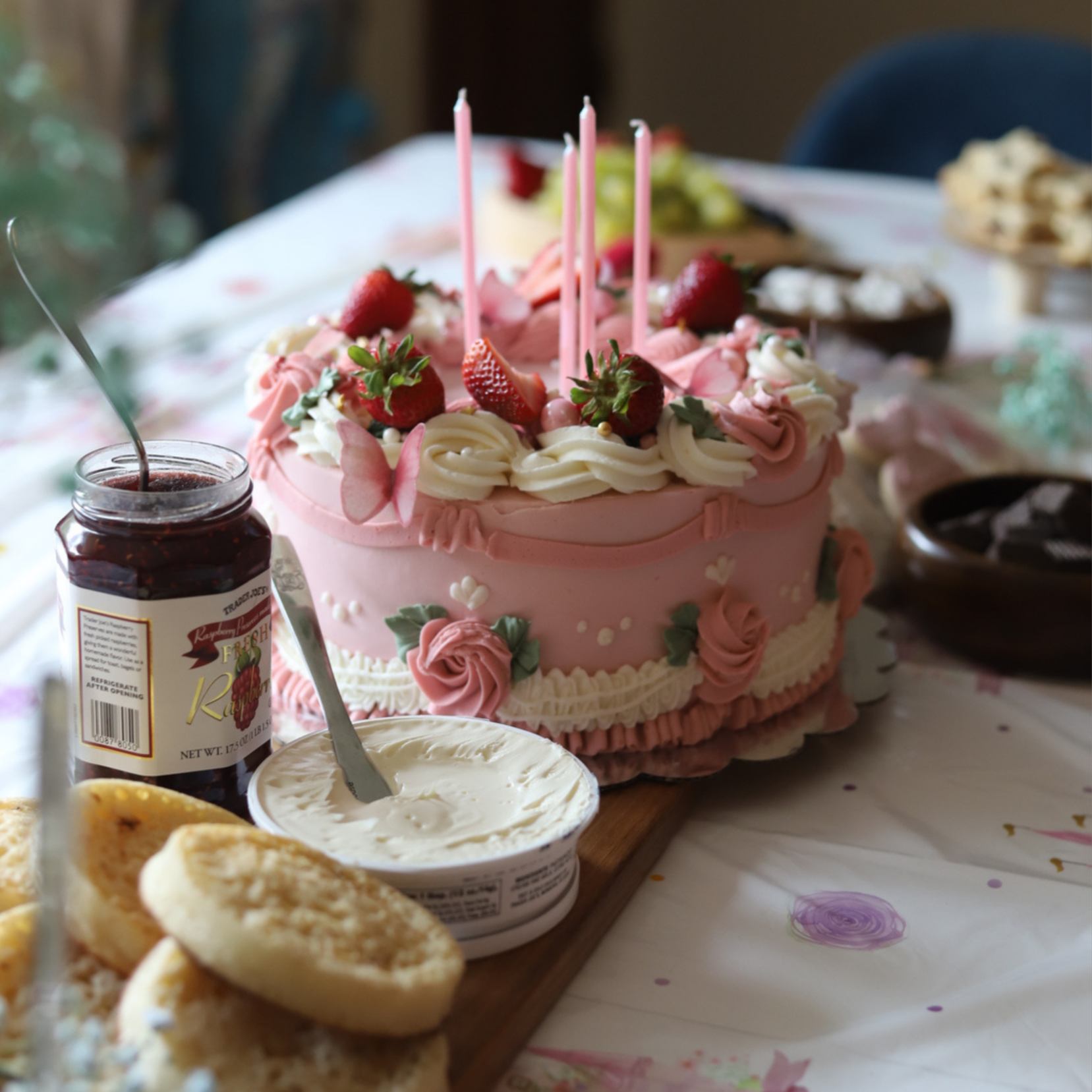 A pink birthday cake decorated with strawberries, small pink candles, and floral icing accents, surrounded by various desserts on a table.