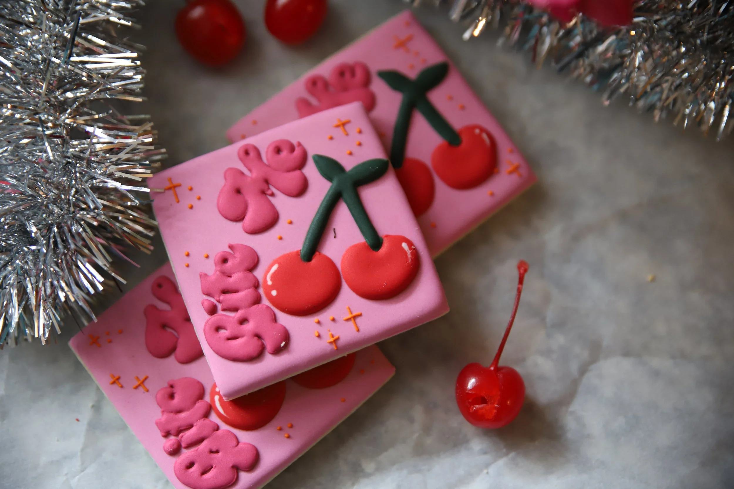 Close-up of pink cookies decorated with red cherries, green leaves, and pink lettering that says "BITE ME", with silver tinsel on the side and a red cherry in the foreground.