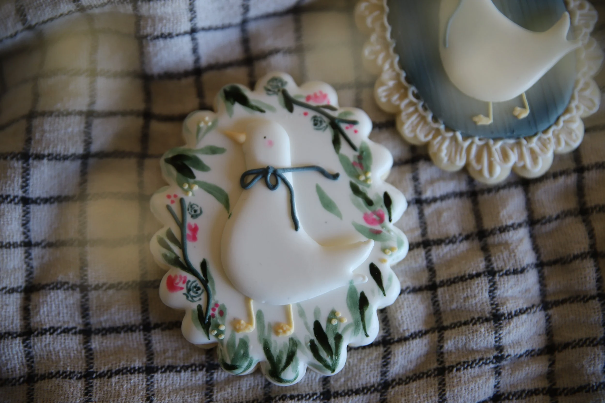 Decorative cookies, one shaped like a goose with floral patterns hand-painted on it on a plaid cloth. Flowers can be visible in the foreground