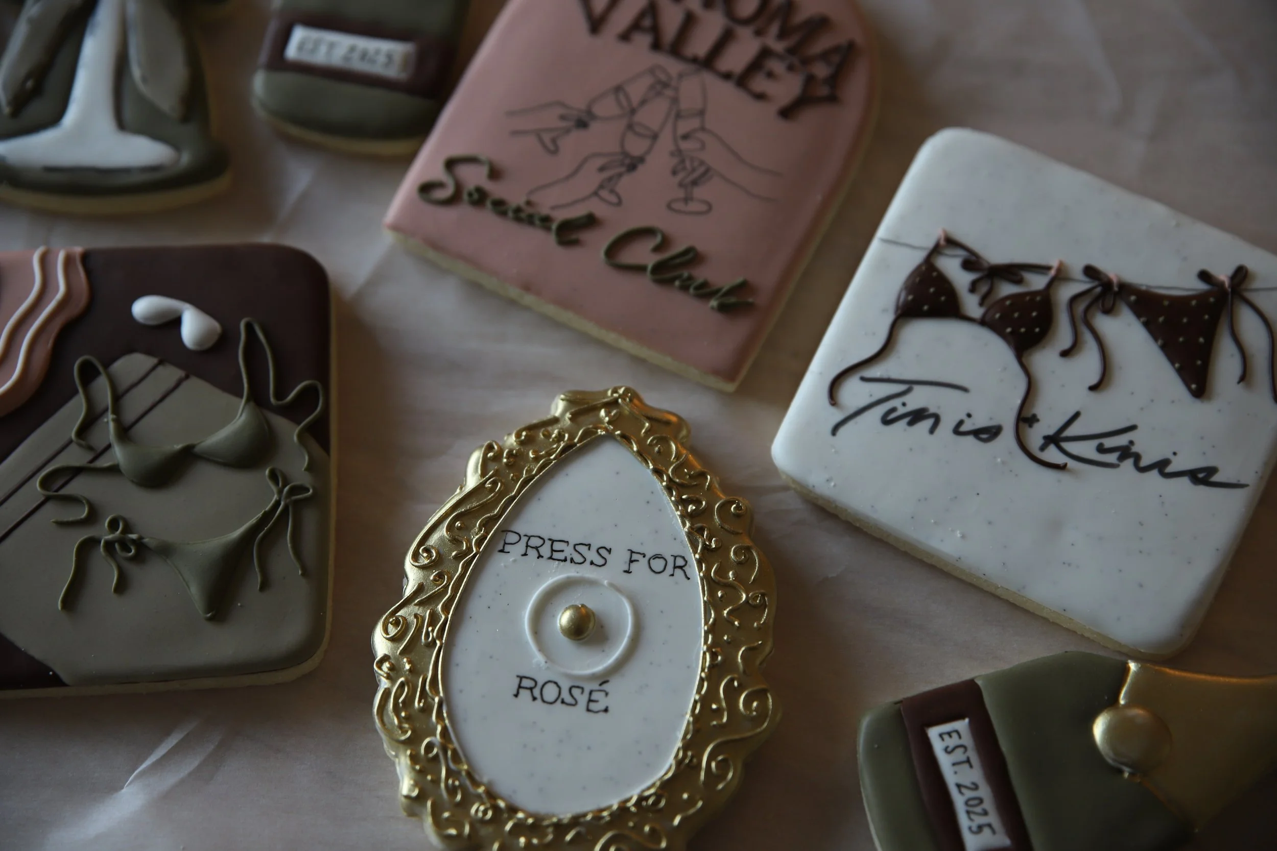 Decorative cookies with gold, white, pink, and brown icing, some with writing like "PRESS FOR ROSE" and "Tinis and Kinis" and others with floral or bikini designs, arranged on a table.