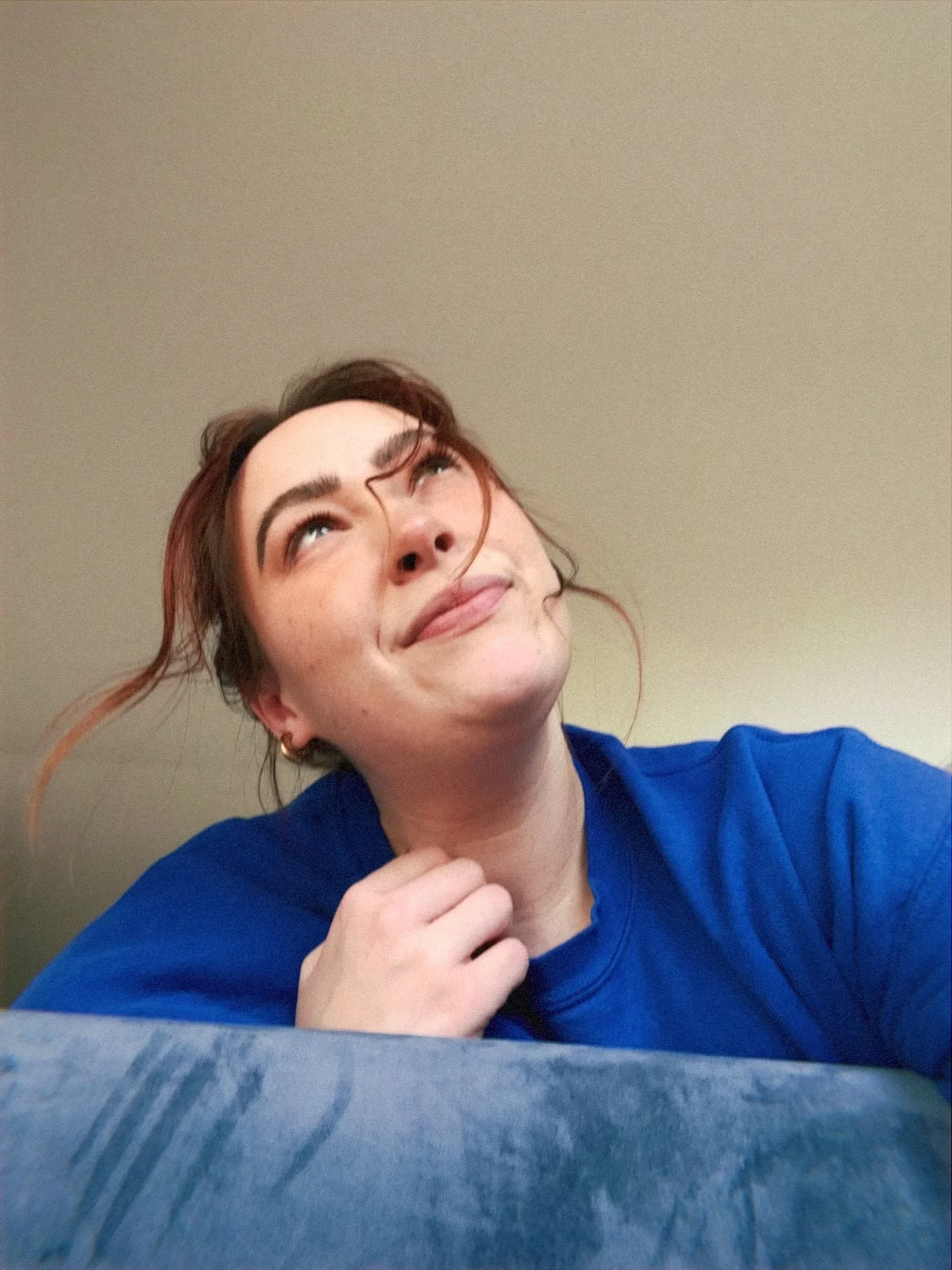 A young woman with brown hair, styled in loose waves, looking up and smiling slightly, wearing a blue shirt. She appears to be sitting at the edge of a velvet couch.