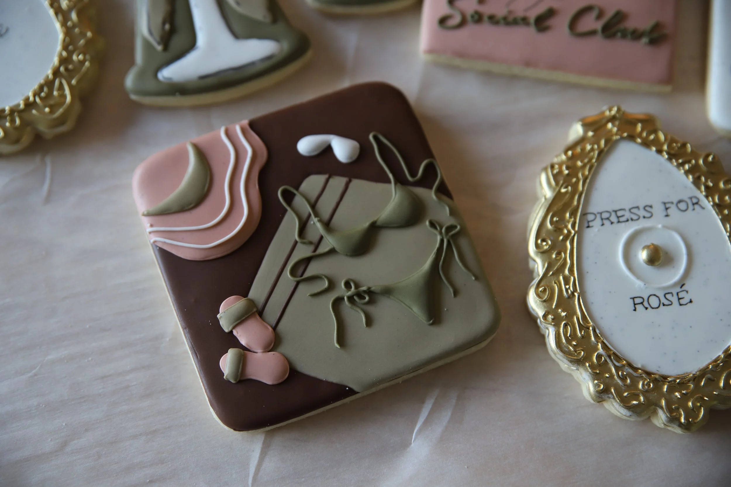 Decorated cookies on a table, including a beach hat, beach shoes, and a small bikini alongside a gold framed cookie that has 'Press For Rose' written on it.