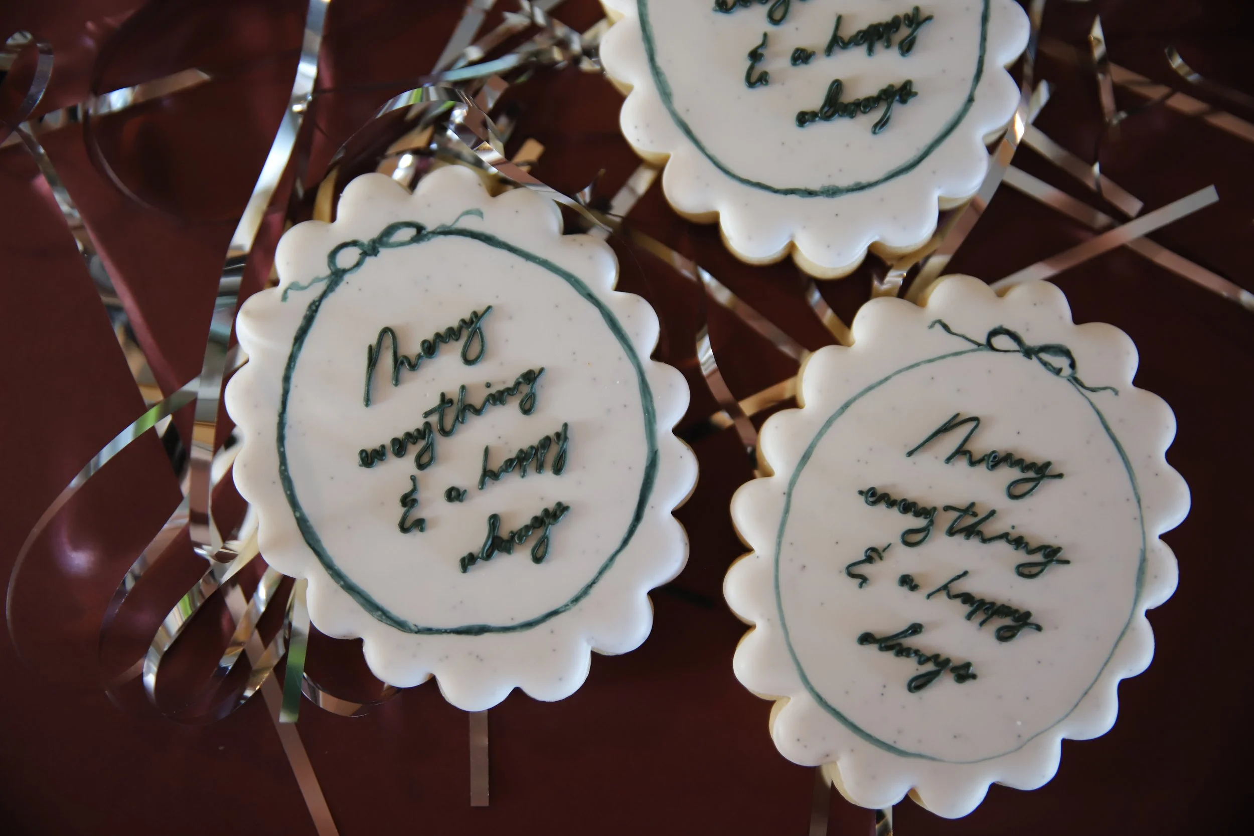 Cookies with white icing and green writing that says "Merry everything & a happy always" on a dark red surface with silver ribbons.