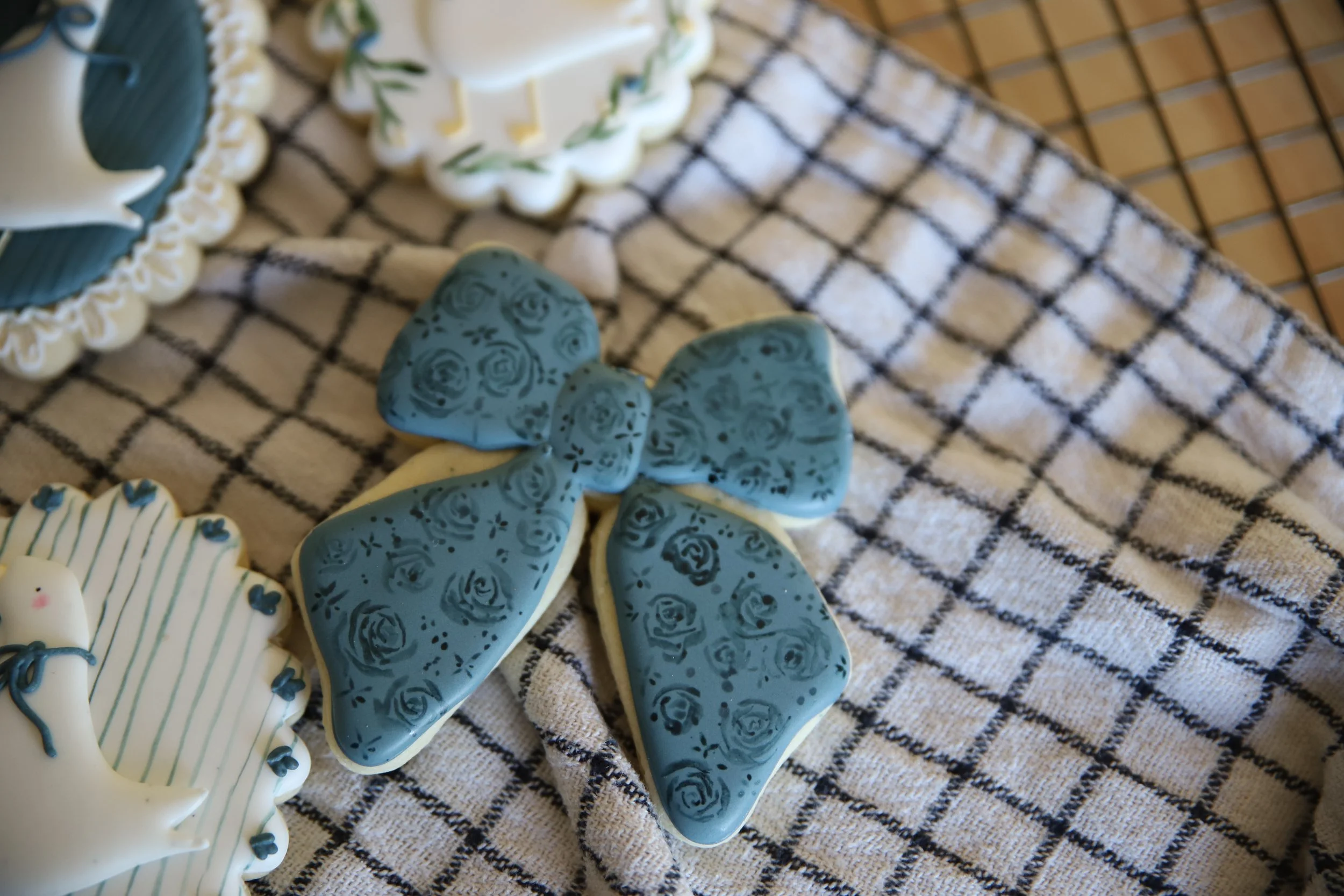 Decorative cookies on a checkered cloth, including a blue bow-shaped cookie with a floral pattern hand painted and a white cookie with blue and green accents, possibly a snowflake or flower design.
