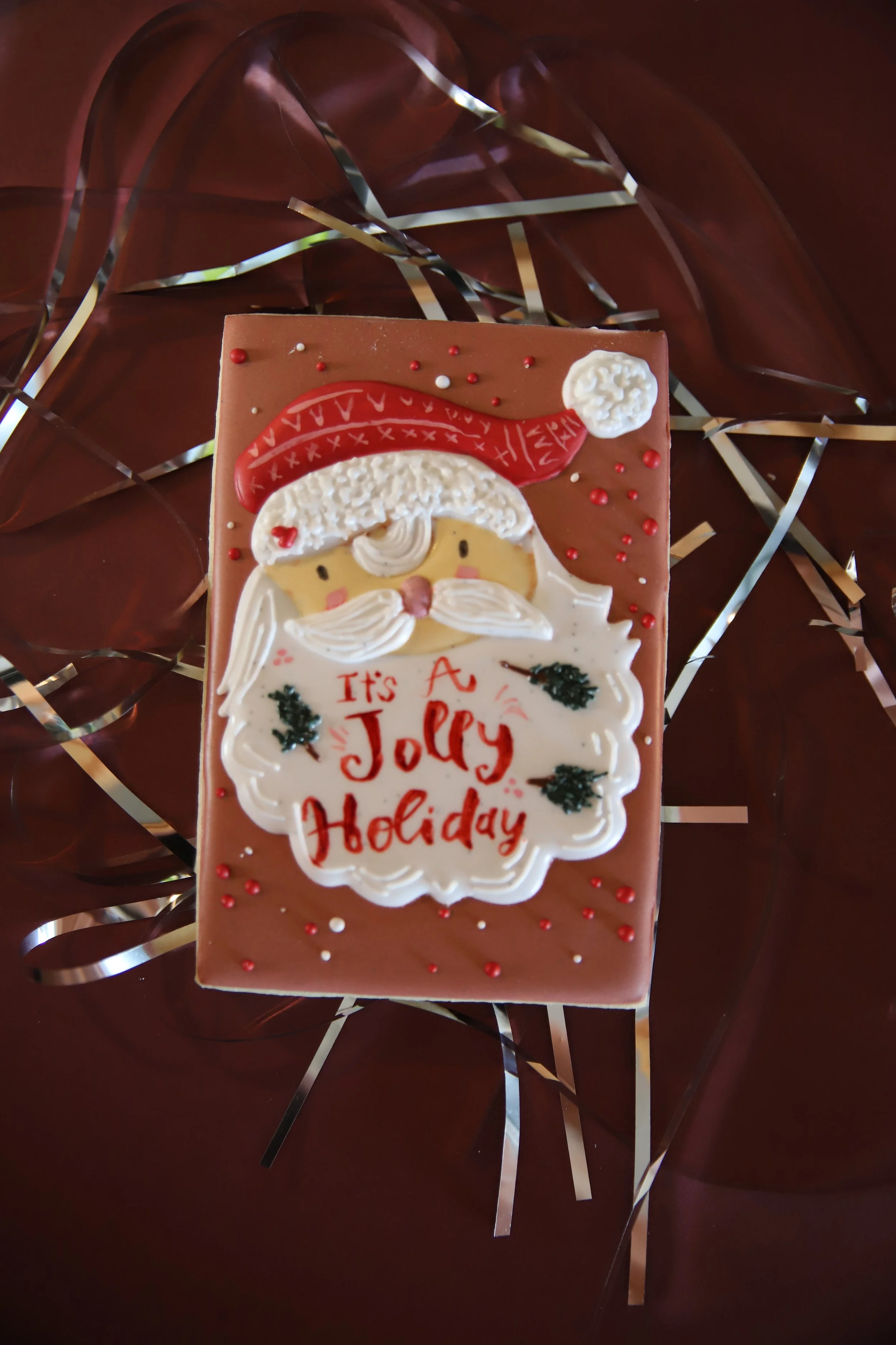 A decorated Christmas-themed cookie with Santa Claus's face, hat, and beard, surrounded by red and white sprinkles, with the words "It's A Jolly Holiday" written on it, placed on a dark, shiny surface with metallic ribbon decorations around.