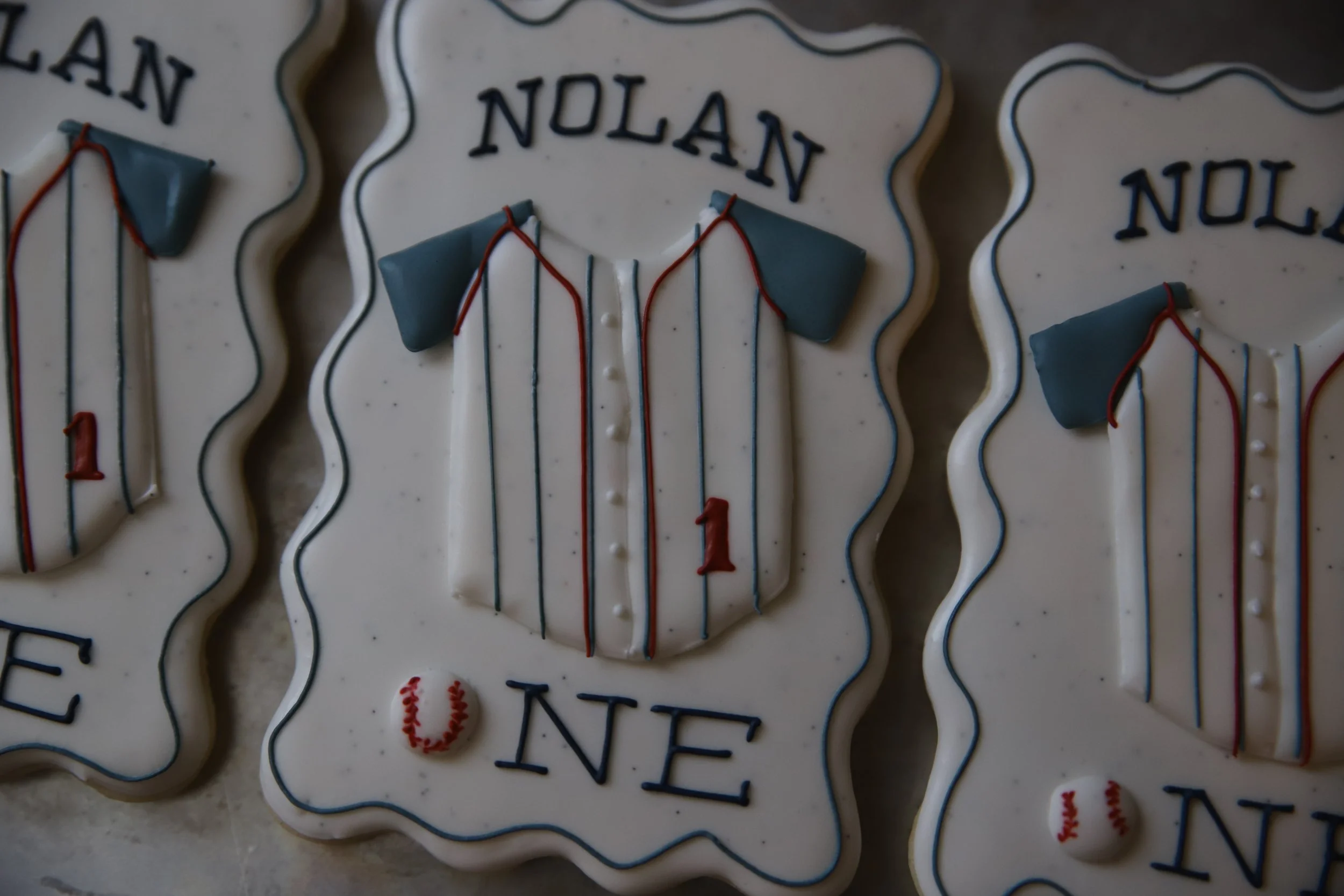 Decorative cookies shaped like jerseys with the name 'NOLAN' on top. The cookies are white with blue and red icing accents.