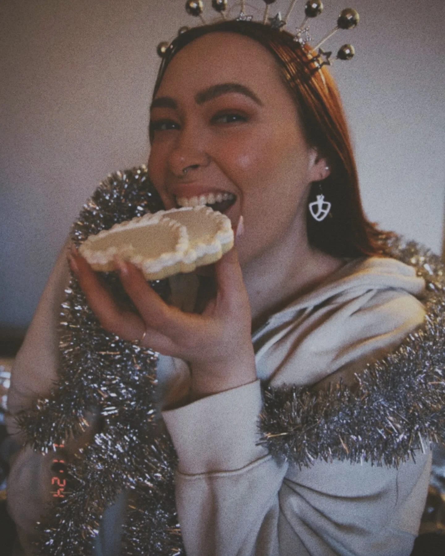 A woman with red hair wearing a disco-themed headband and miniature mixing bowl paddle earrings, holding and about to eat a decorated sugar cookie. She's wrapped in a silver tinsel boa.