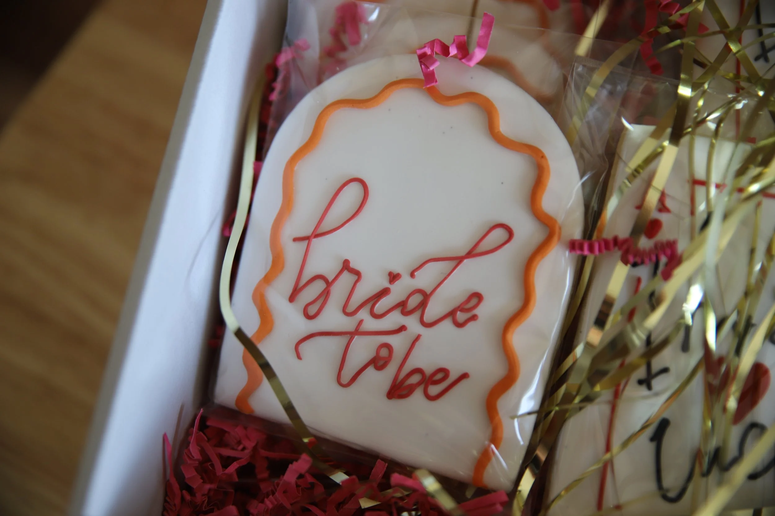 Decorative cookie with orange and red icing spelling 'bride to be' inside a gift box filled with pink and gold shredded paper.